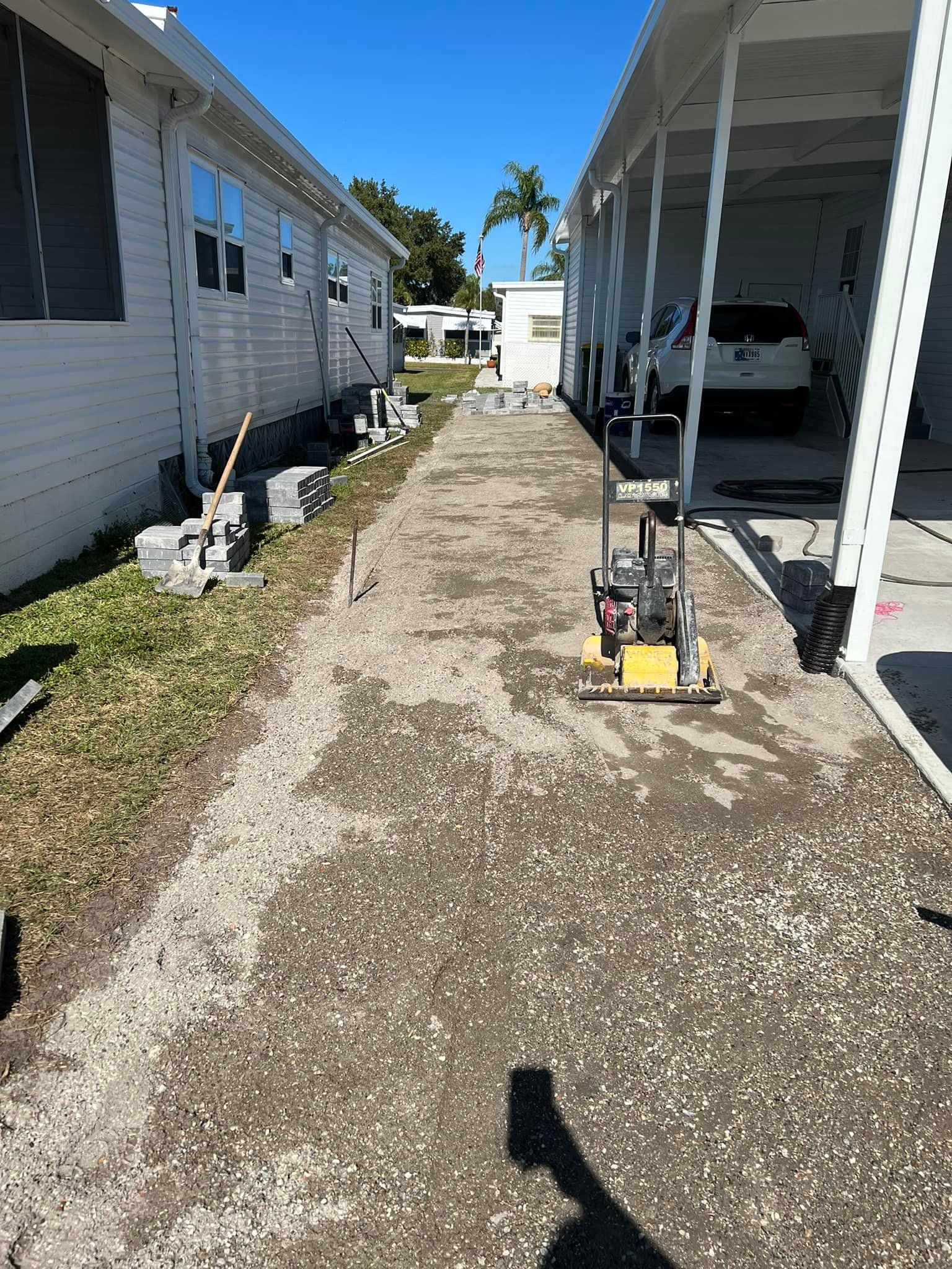 Driveway construction between two white buildings. A vibrating plate compactor sits on gravel.