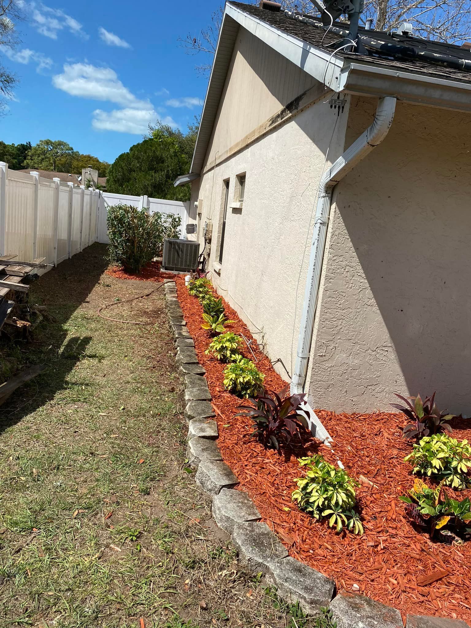 Side yard with mulch beds, shrubs along a stucco house, white fence, and blue sky.