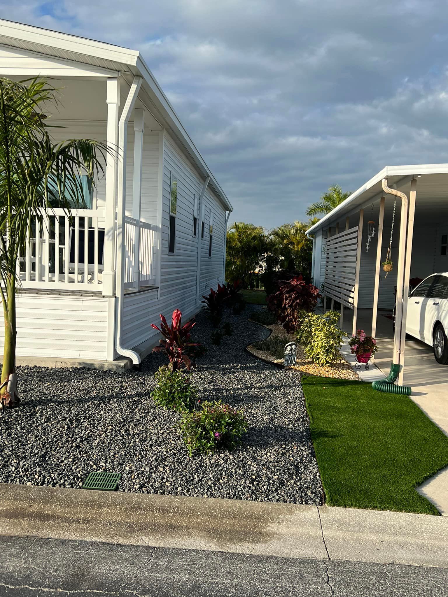 A narrow walkway between two white buildings with landscaping. The ground has dark gravel, grass, and plants.