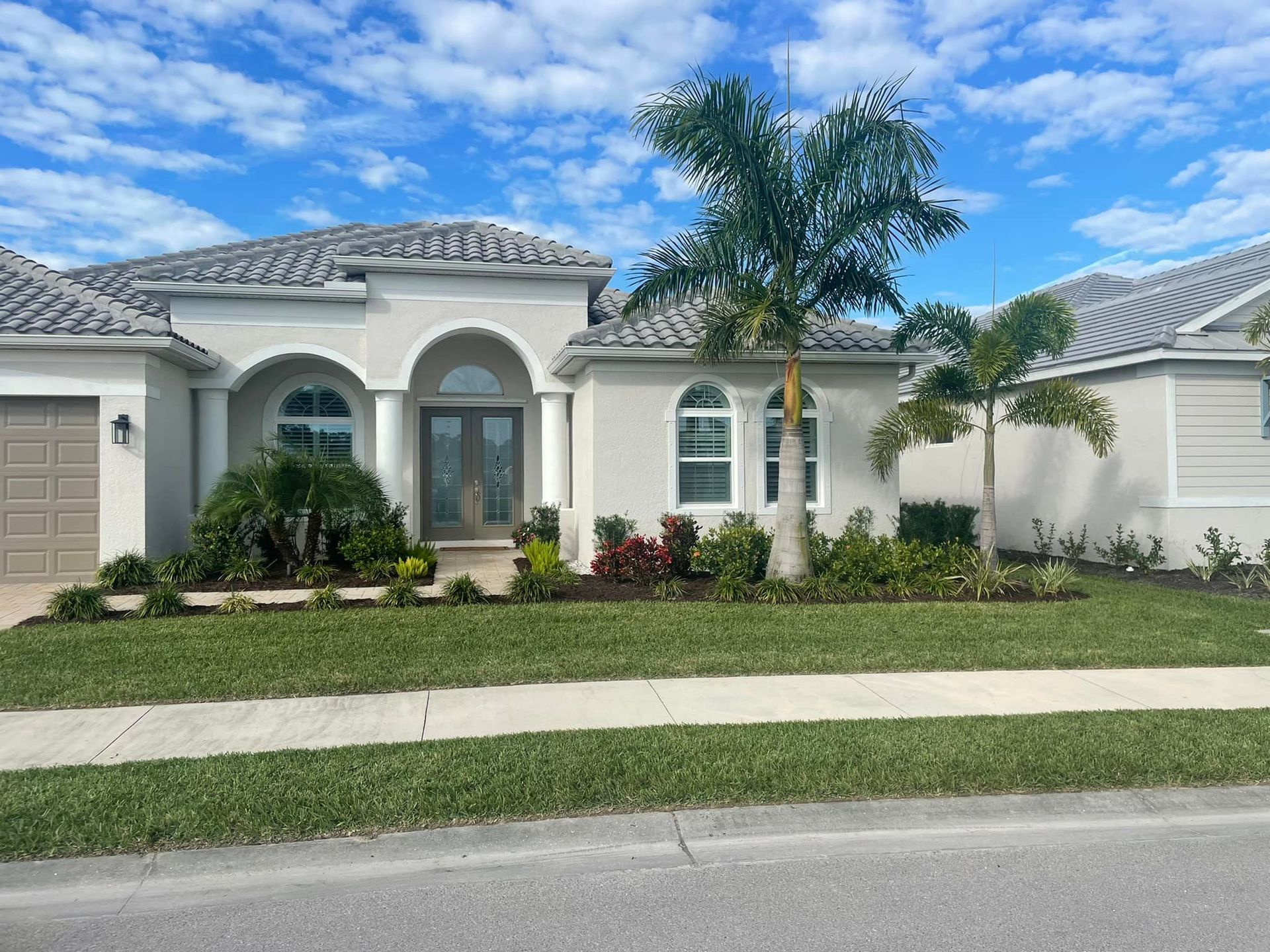 Tan stucco house with tiled roof, green lawn, palm trees, and blue sky.