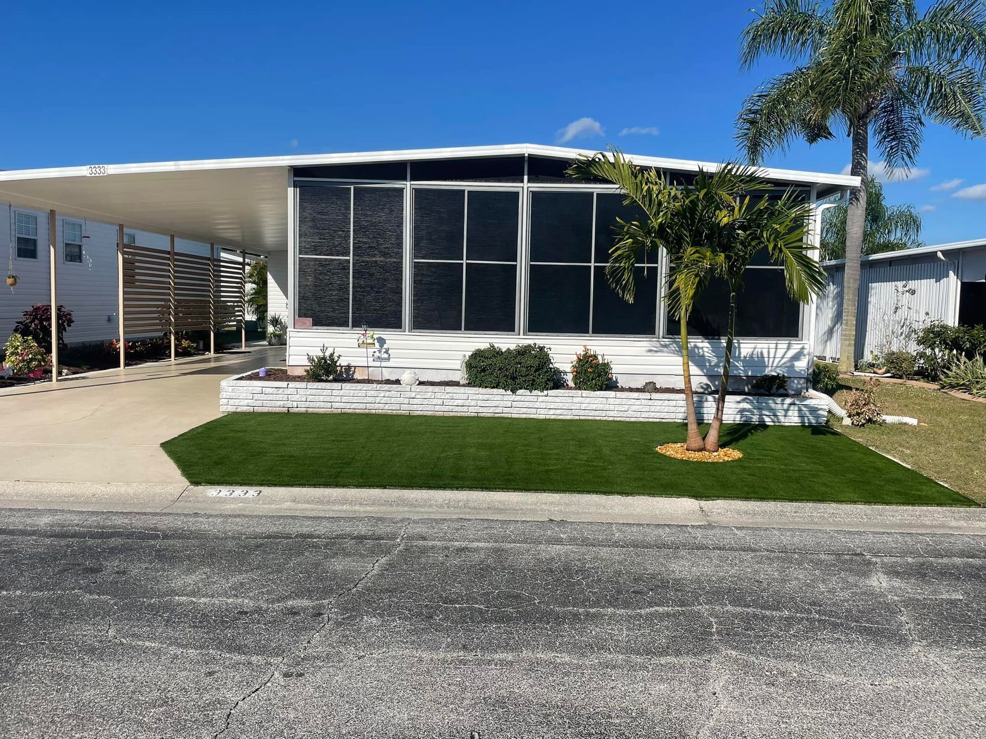 White mobile home with carport and small palm tree in front. Green lawn, blue sky.