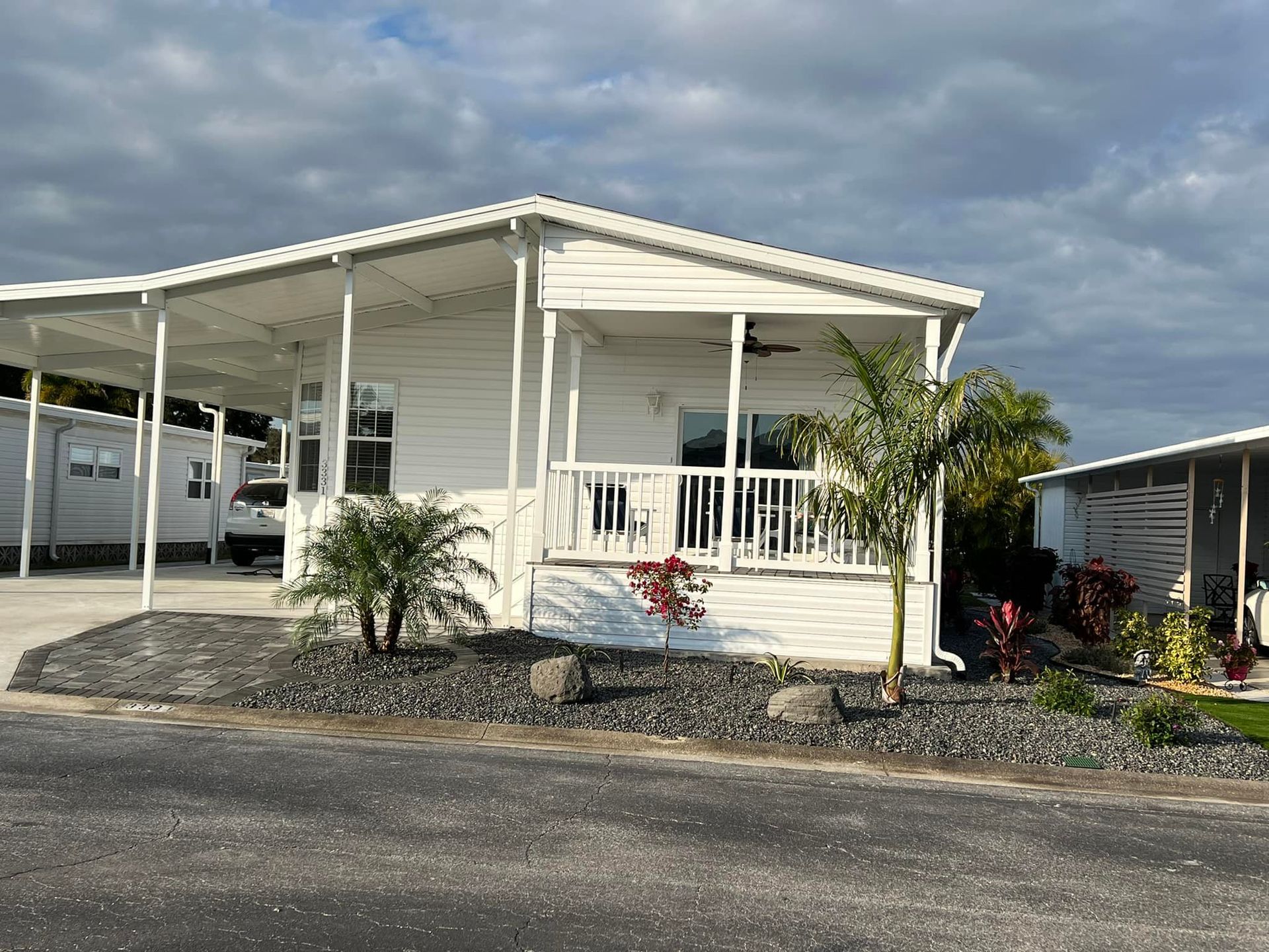 White mobile home with carport, front porch, and palm trees under a cloudy sky.