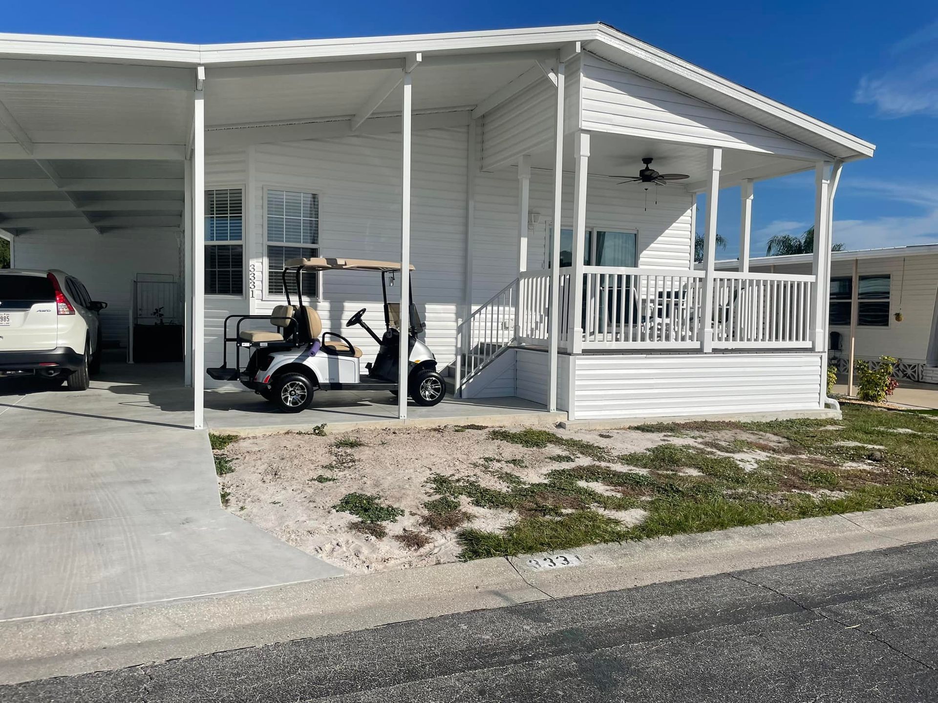 White mobile home with carport, golf cart parked, front porch with railing, blue sky.