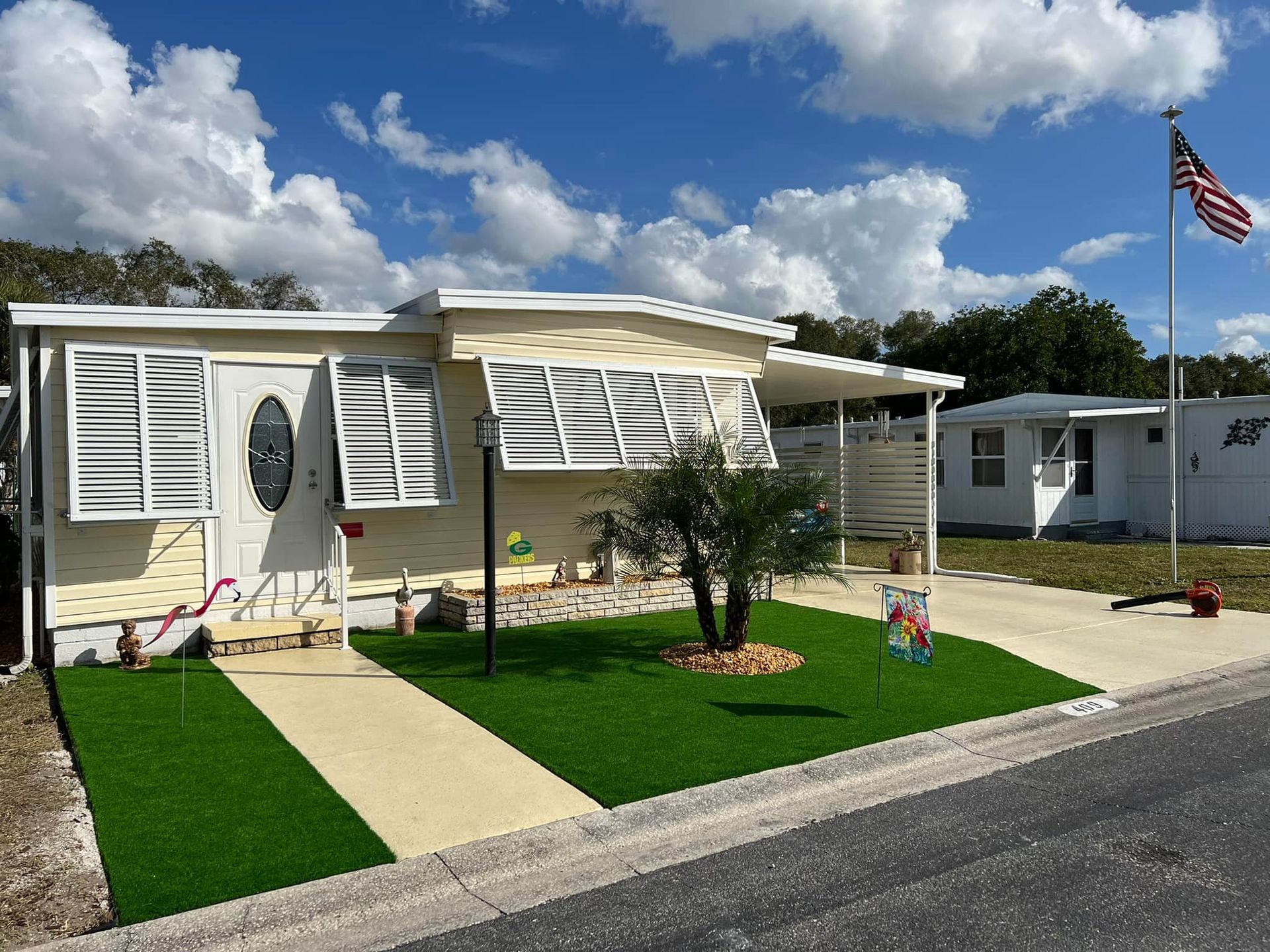 A single-story mobile home with beige siding, shutters, and a small yard with artificial grass.