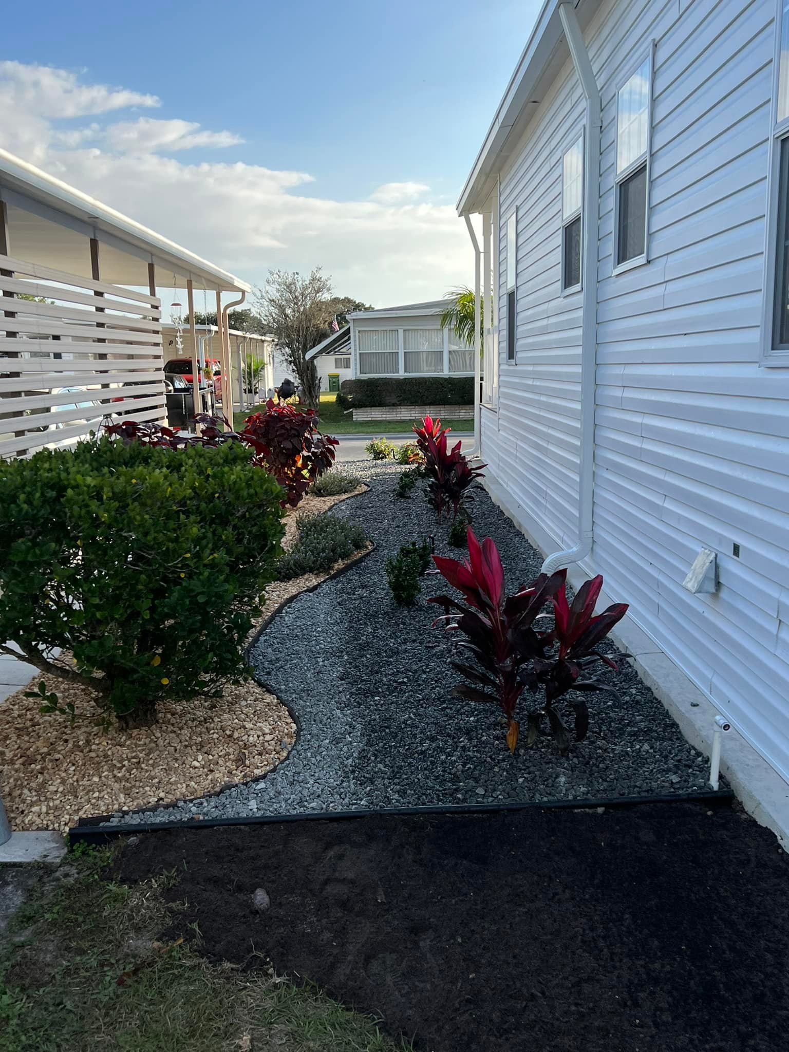 Landscaped garden bed with dark grey rock, red plants, and green shrubs between two white buildings.