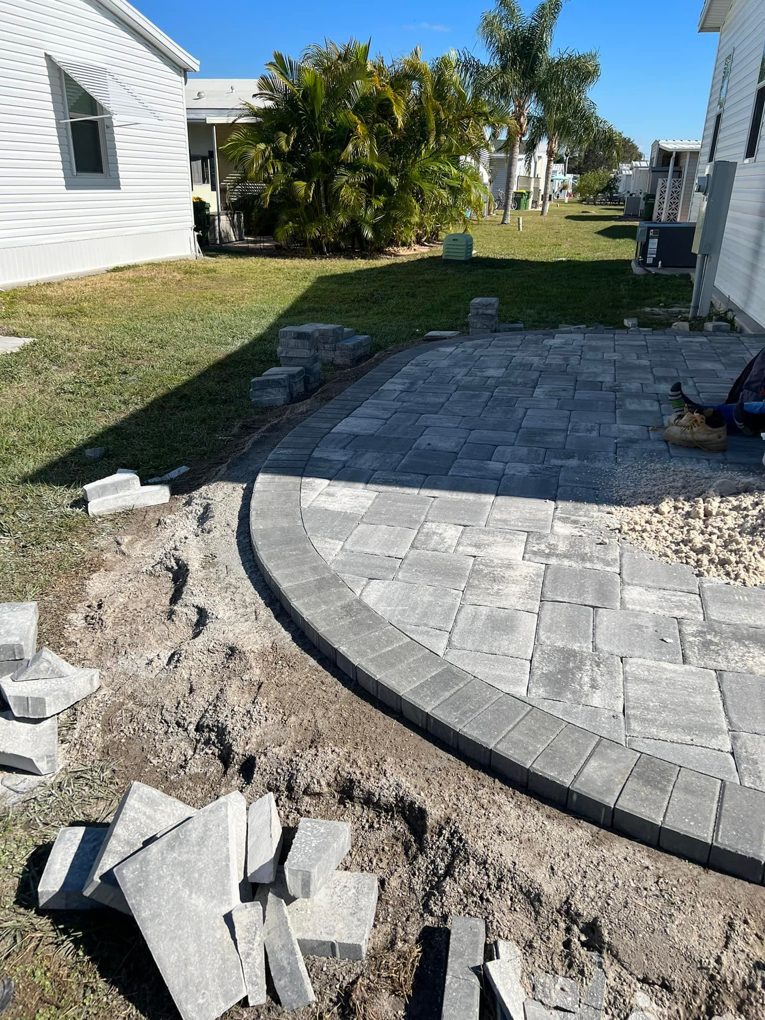 A patio being constructed with gray pavers and border bricks, in a grassy yard, beside a house.
