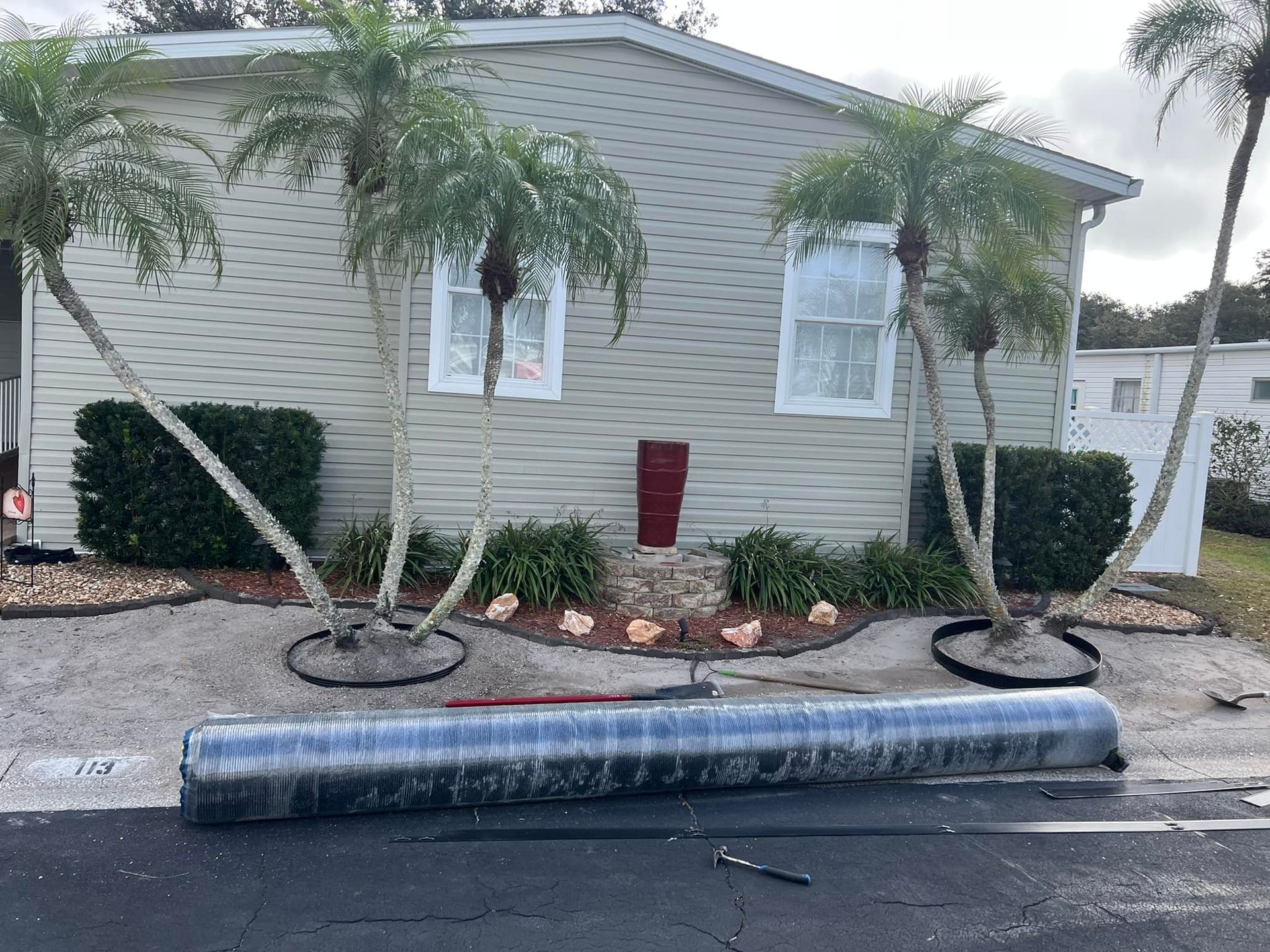 Mobile home with palm trees and a rolled-up object in front. Gray siding, tan gravel, blue sky.