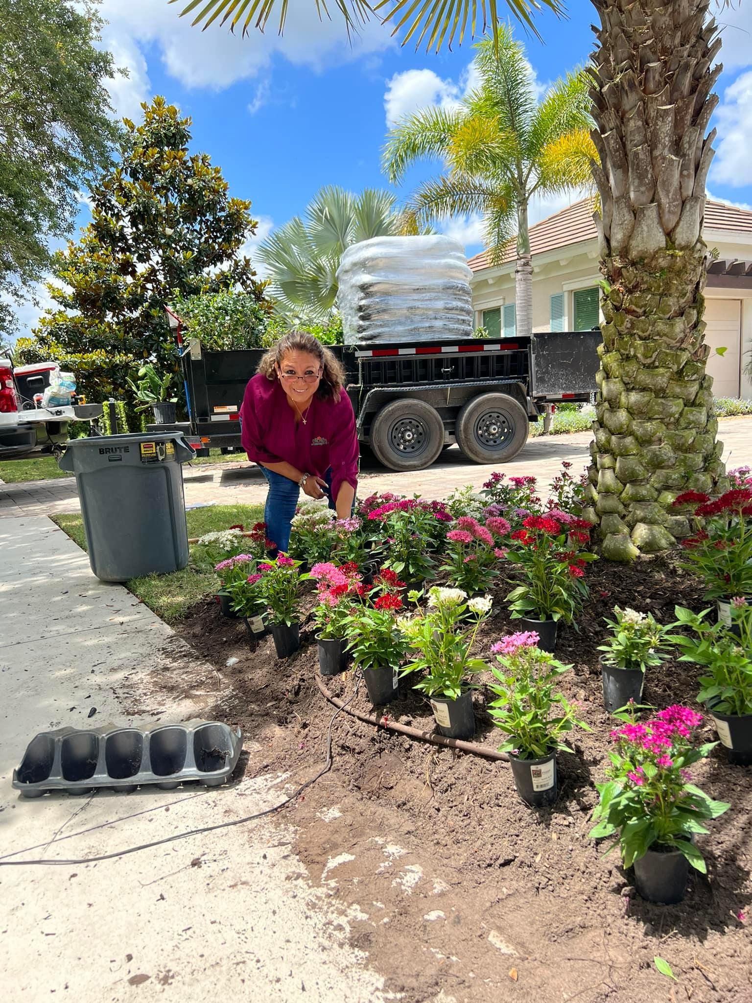 Woman planting flowers in a garden bed next to a sidewalk; trailer with bags of soil in the background.
