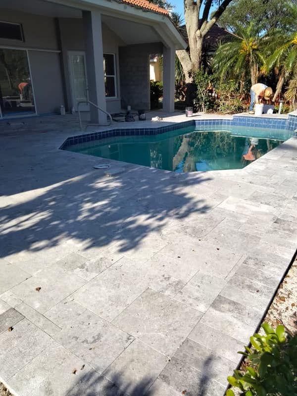 Swimming pool with light gray stone patio. House in background, blue tiles around pool.