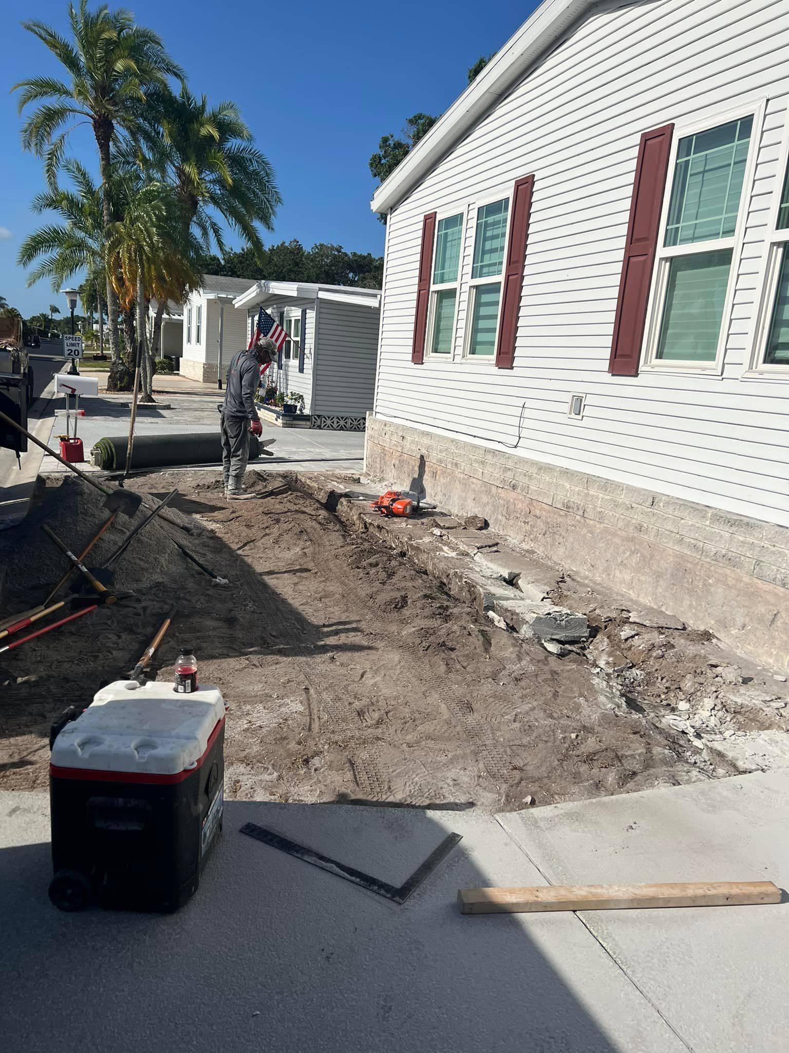 Construction site: Dirt patch near a white house with brown shutters. Cooler and tools sit on the pavement.