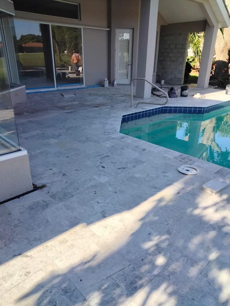 Poolside patio with travertine pavers next to a swimming pool. Sunlight casts shadows.