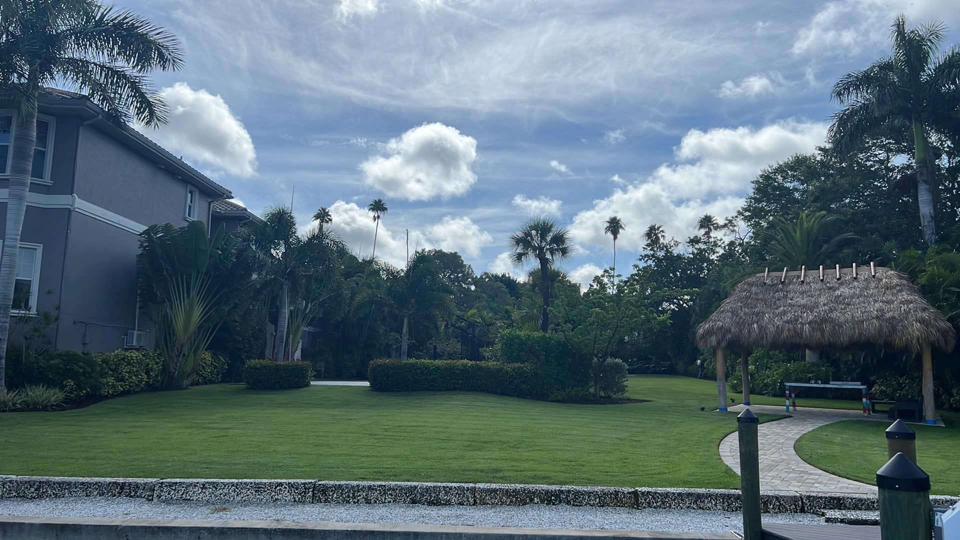 Lush green lawn leads to a thatched-roof shelter and dense trees under a cloudy sky, next to a large building.