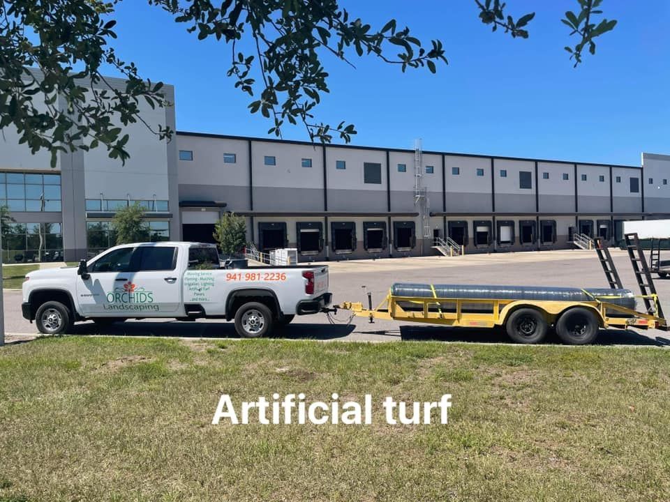 White truck with trailer carrying artificial turf parked in front of a commercial building.