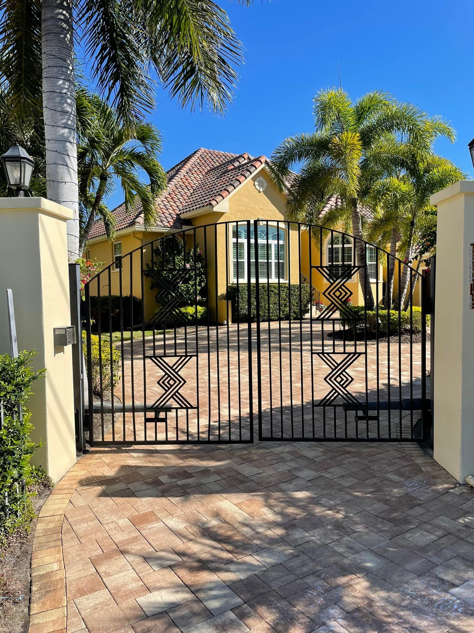 Black metal gate in front of a yellow house with a red tile roof and palm trees.