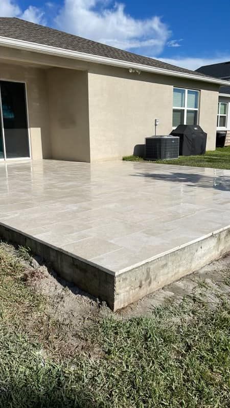 Beige tiled patio adjacent to a light brown stucco house; sunny outdoor setting.