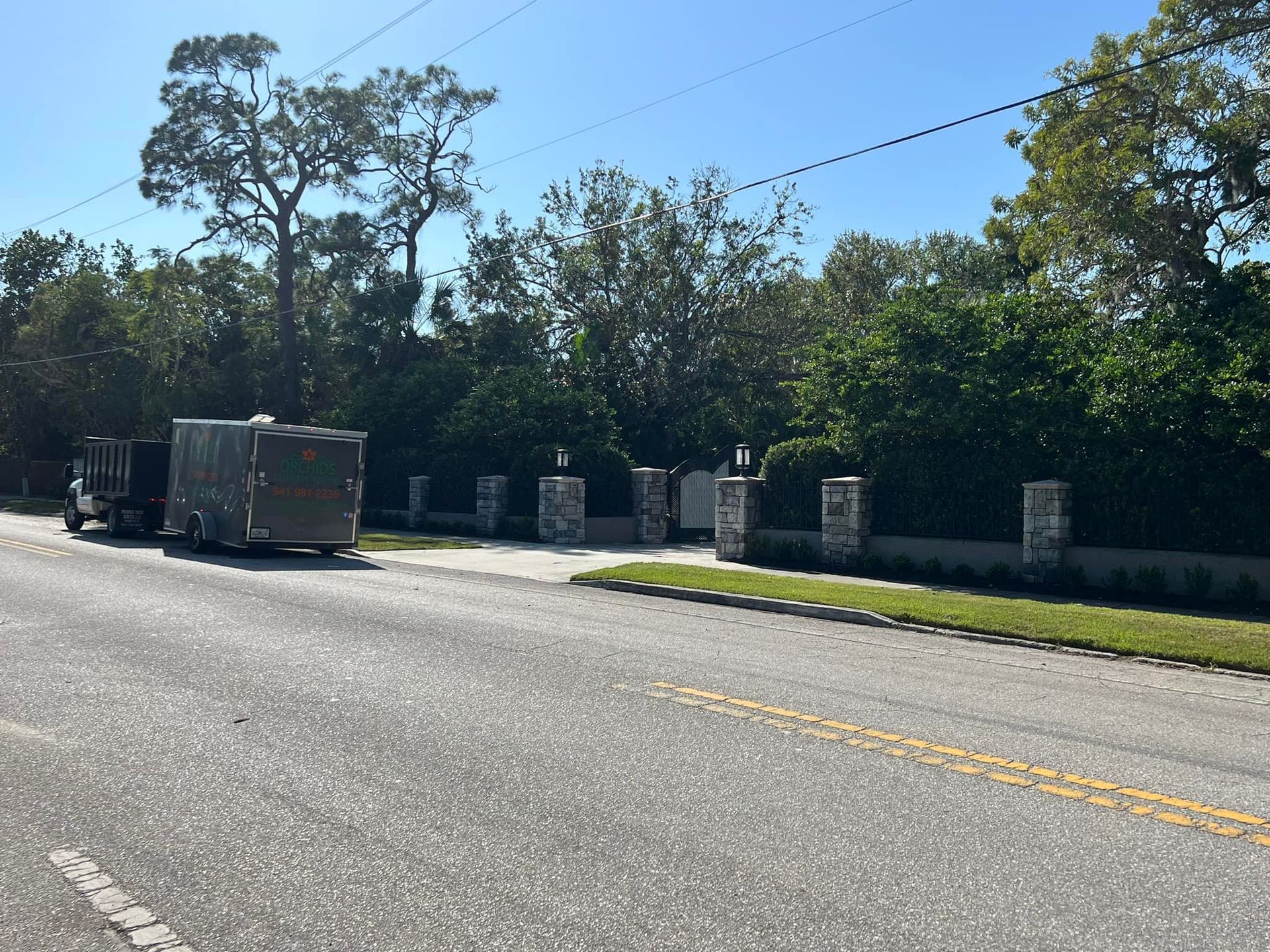 Roadside view of a gated entrance with stone columns and lush greenery; truck with trailer on the left.