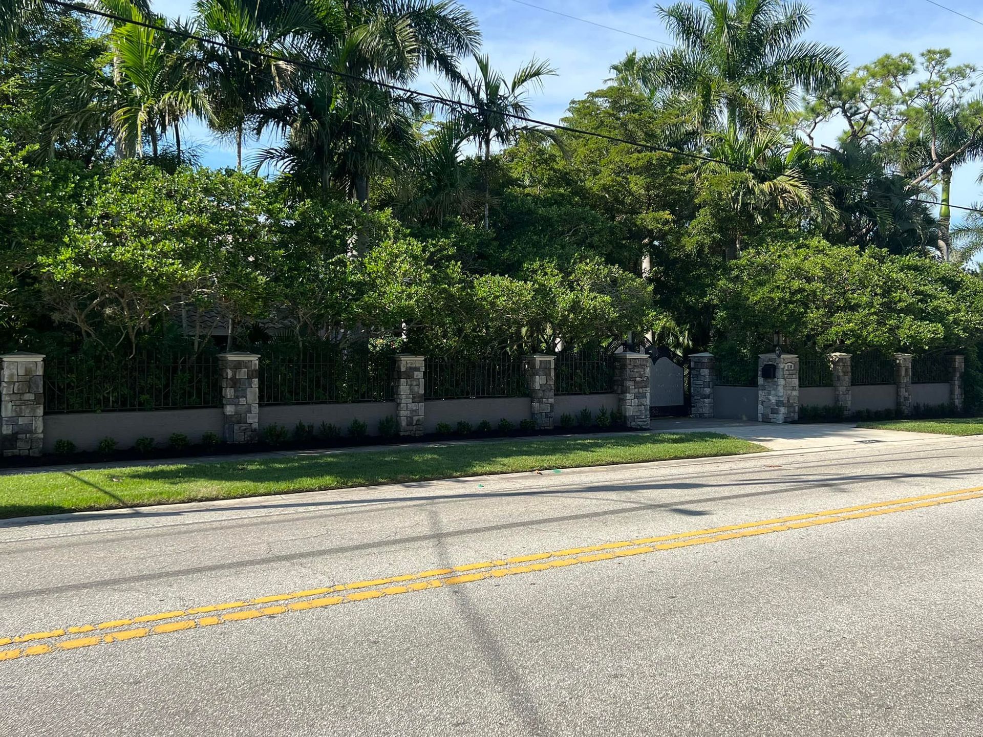 A stone pillar and black fence surround a well-landscaped yard, next to a street with double yellow lines.