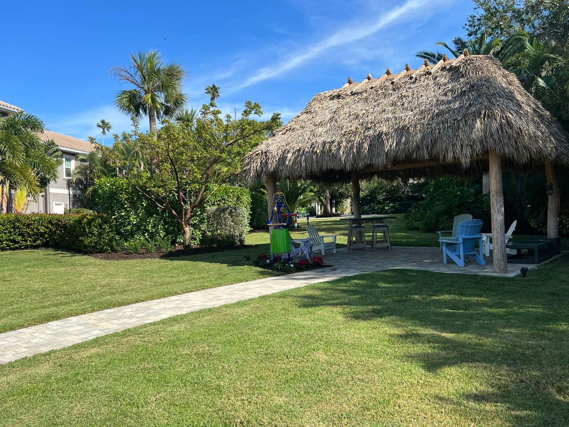 A thatched-roof structure with seating on a grassy lawn. Path leads up to it. Green foliage and buildings in the background.