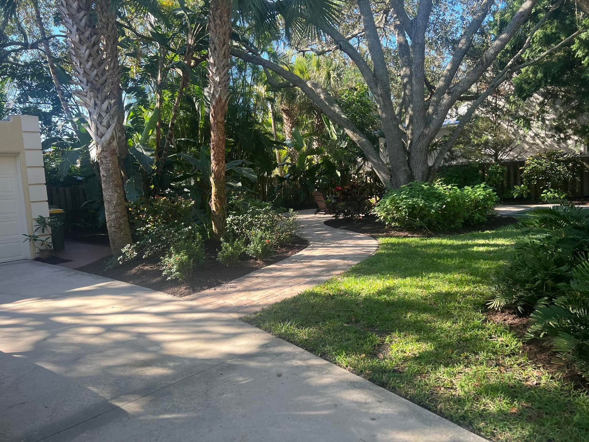 Paved driveway leads to a stone path through lush landscaping, including palm trees and a large shade tree.