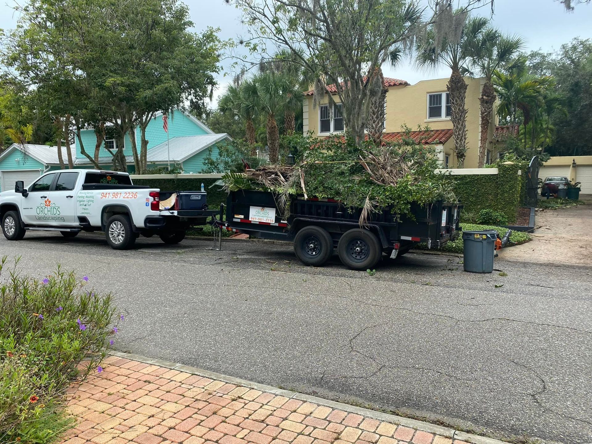 White truck and trailer filled with tree debris parked on a street in front of a house.