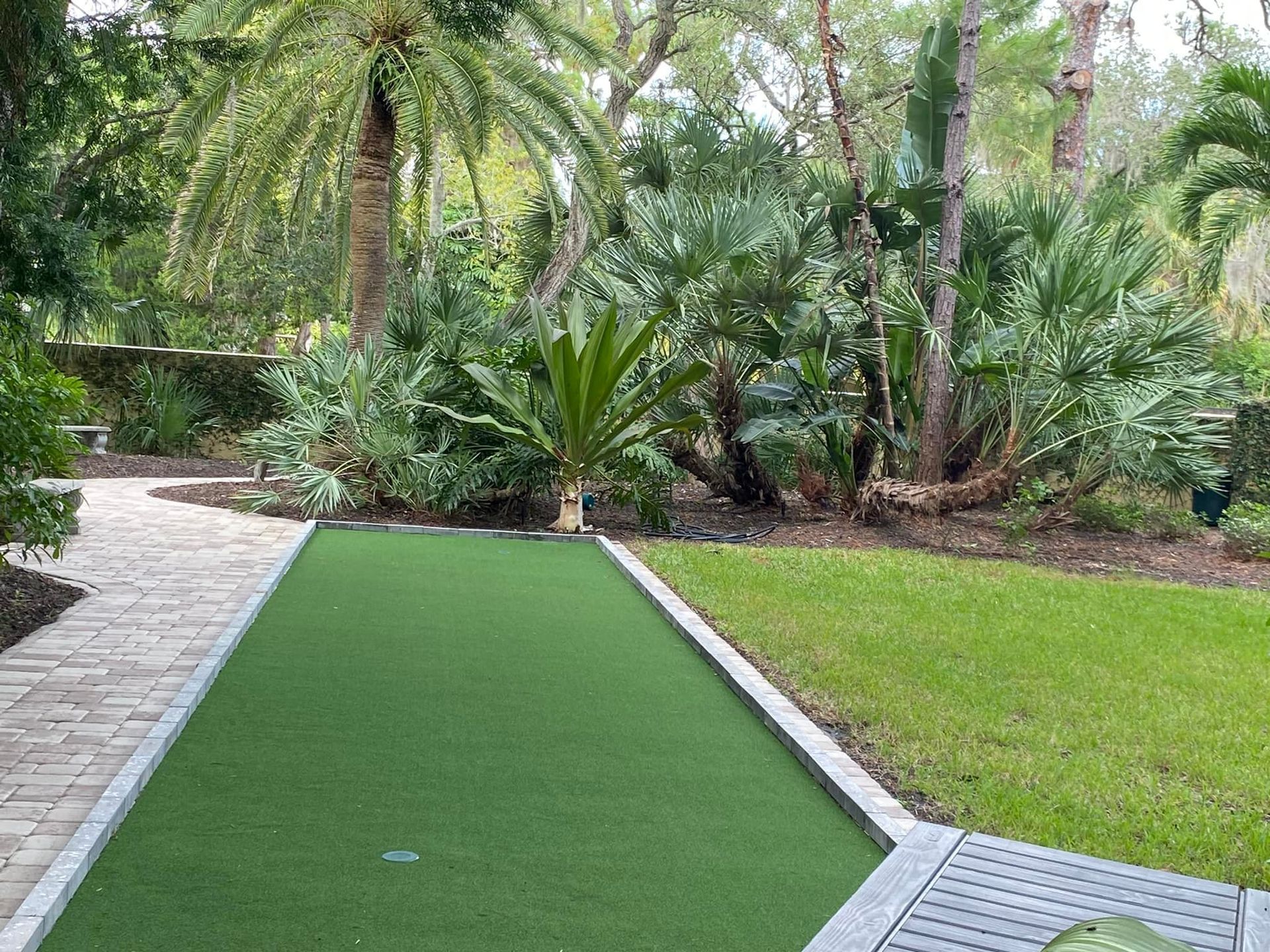 Lush green backyard with a putting green, stone walkway, and tropical plants.