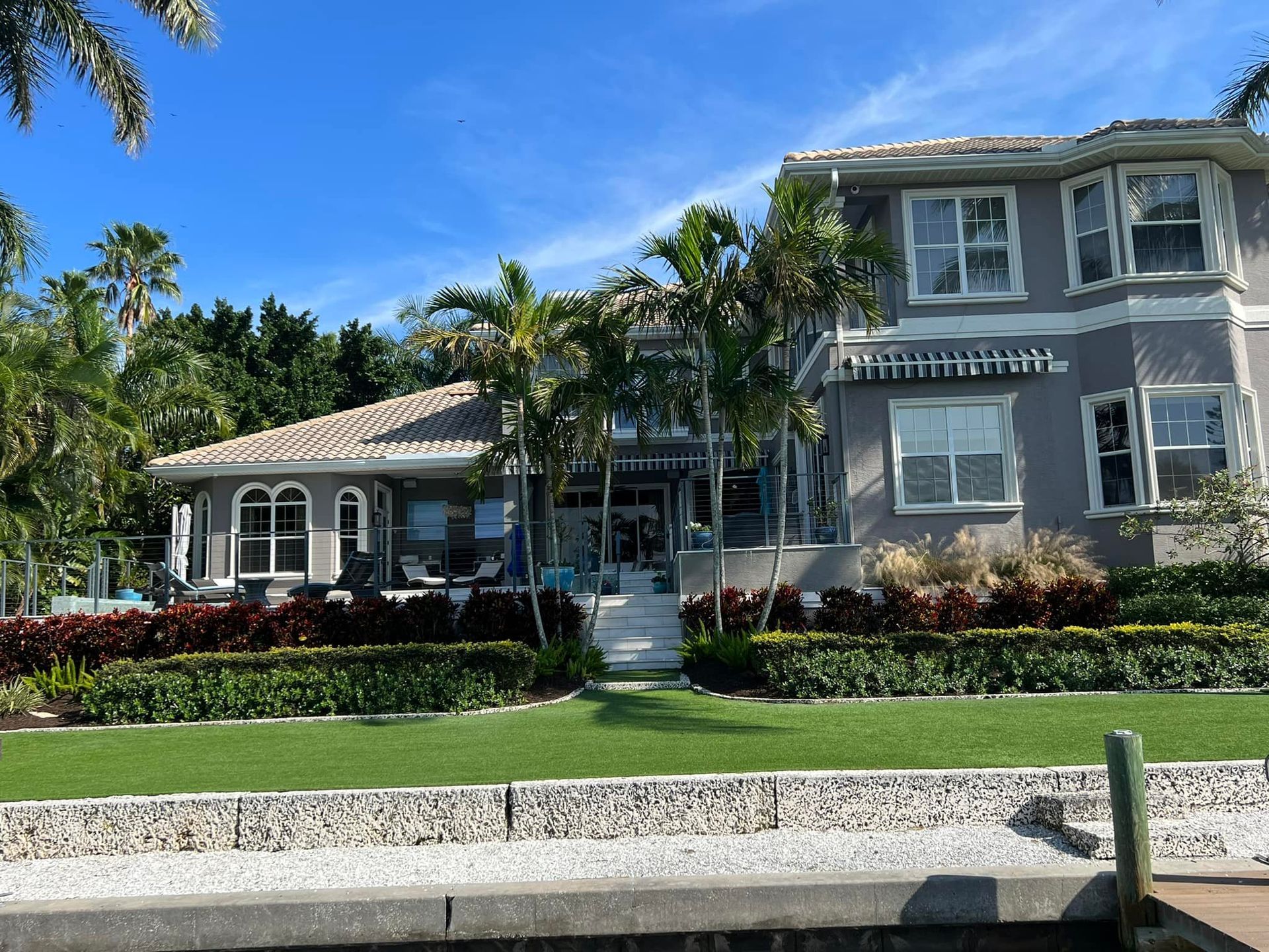 Two-story gray house with white trim, palm trees, and green lawn. Overlooks water under a blue sky.