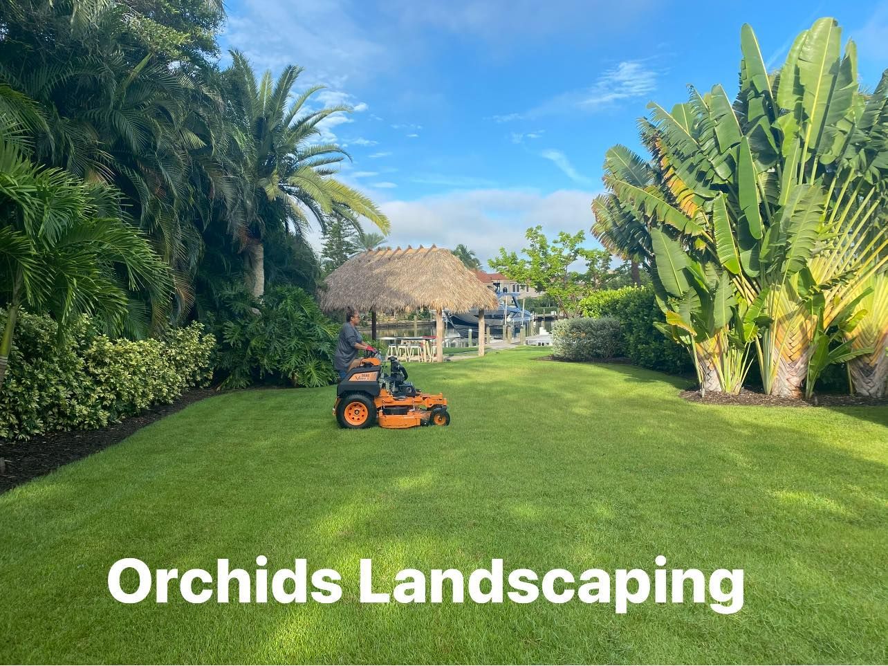 Lawn mower on green grass, with tropical plants and a tiki hut in the background; Orchids Landscaping.