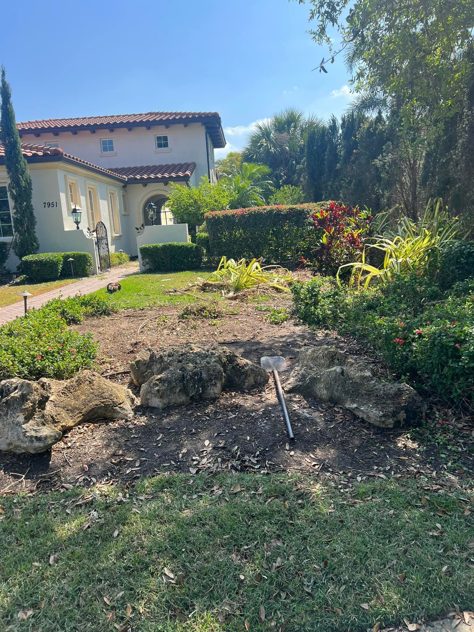 A low stone wall borders a yard in front of a light-colored house with a red-tiled roof.