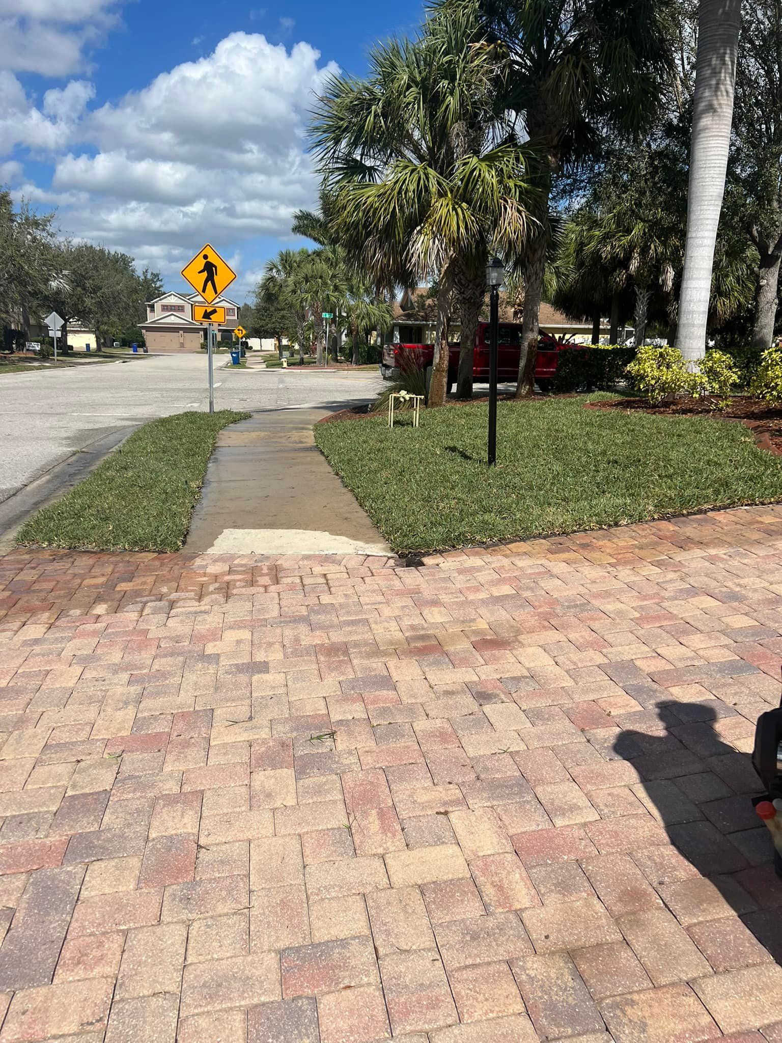 Brick walkway leads to a sidewalk and crosswalk with pedestrian sign, green bushes, and a street view with buildings.
