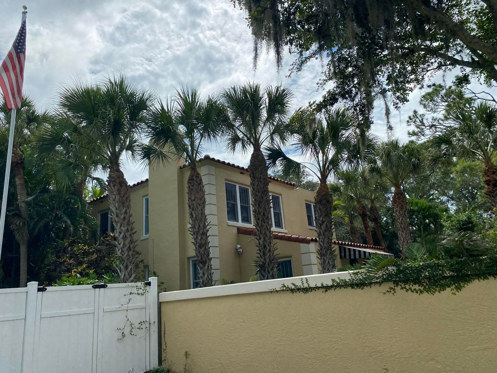 Two-story stucco house with palm trees, American flag, and white fence against a cloudy sky.