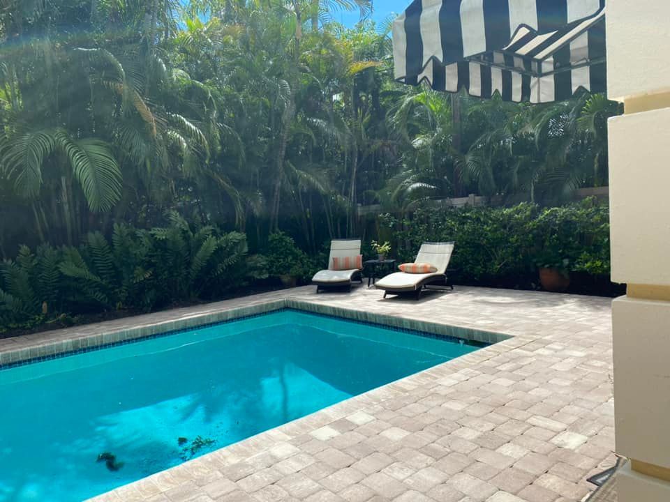 Poolside scene with a blue pool, two lounge chairs, and lush greenery. A striped awning is visible.