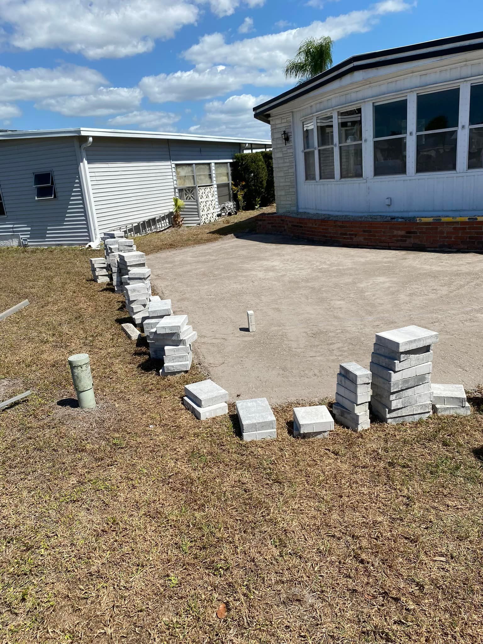 Piles of gray pavers outlining a circular patio on a patch of dead grass, next to a white house under a sunny sky.