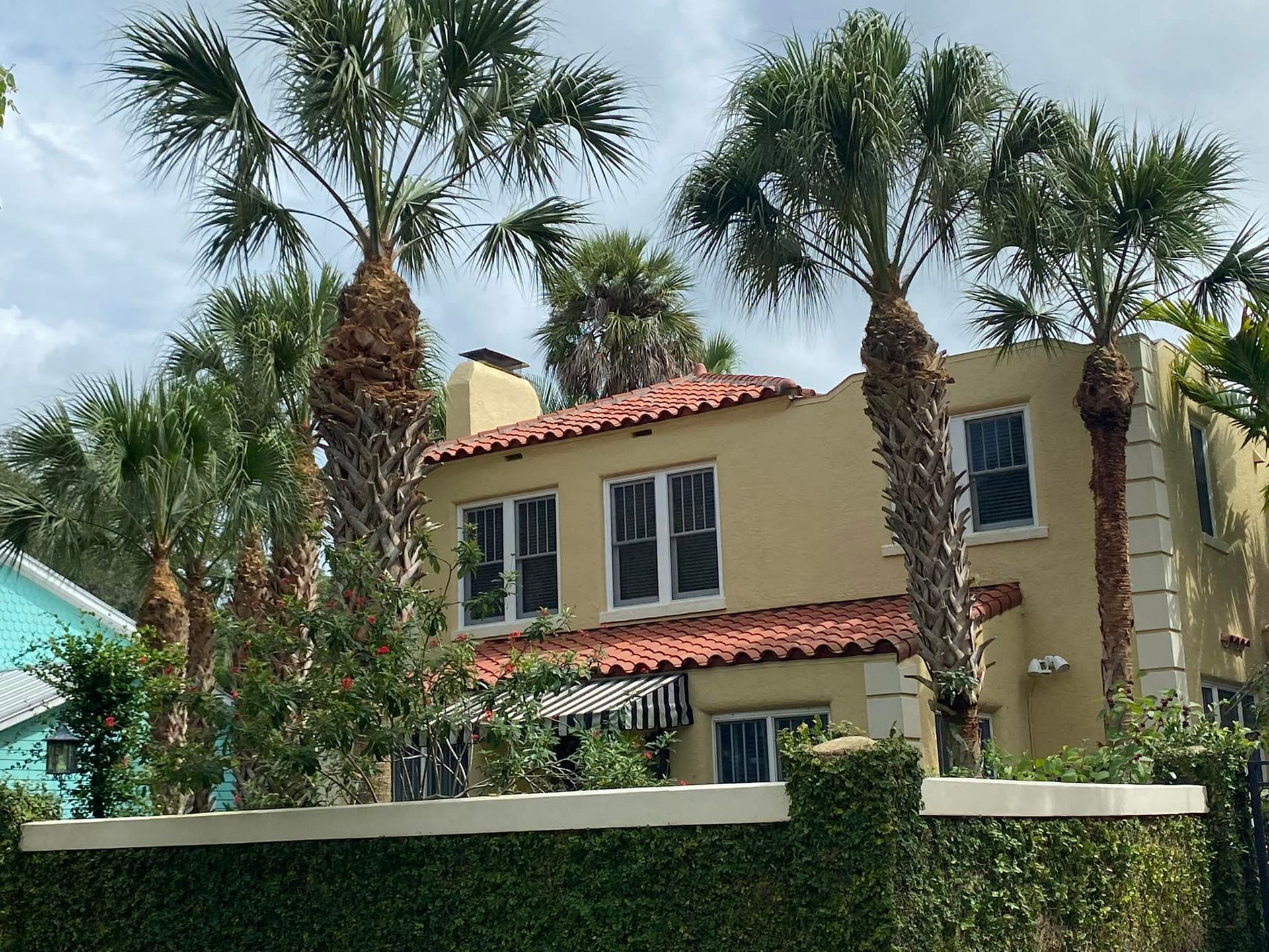 Two-story tan house with red tile roof, palm trees, and a green fence.