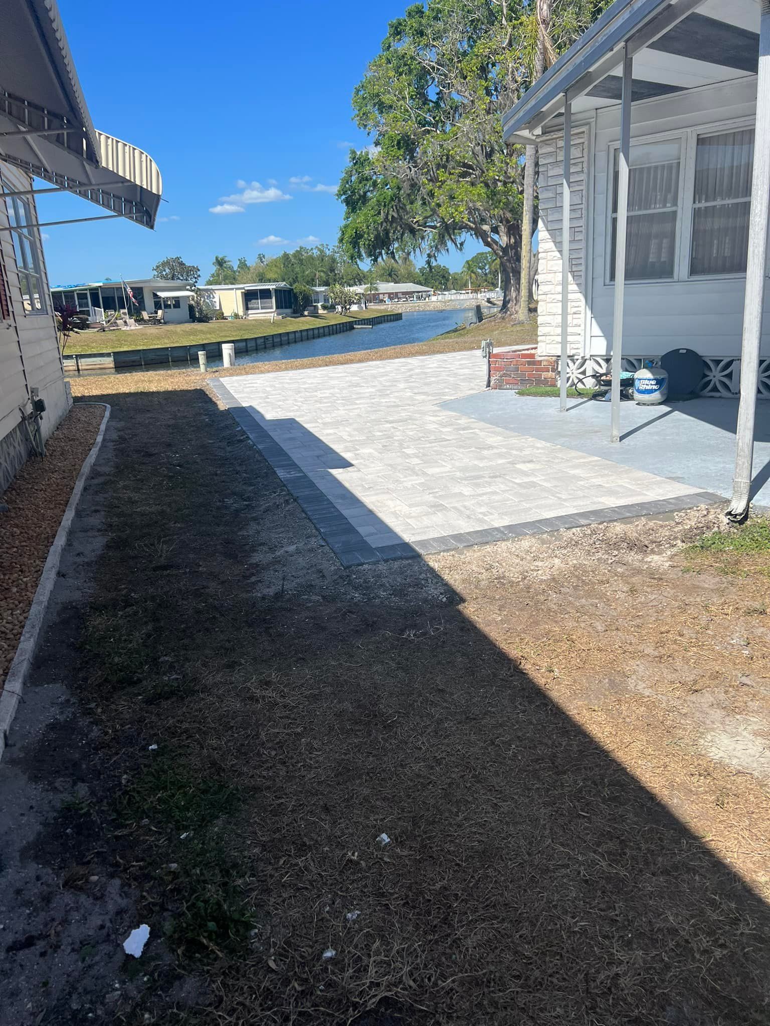 Narrow walkway between two houses, one with a stone patio, leading to a canal under a blue sky.