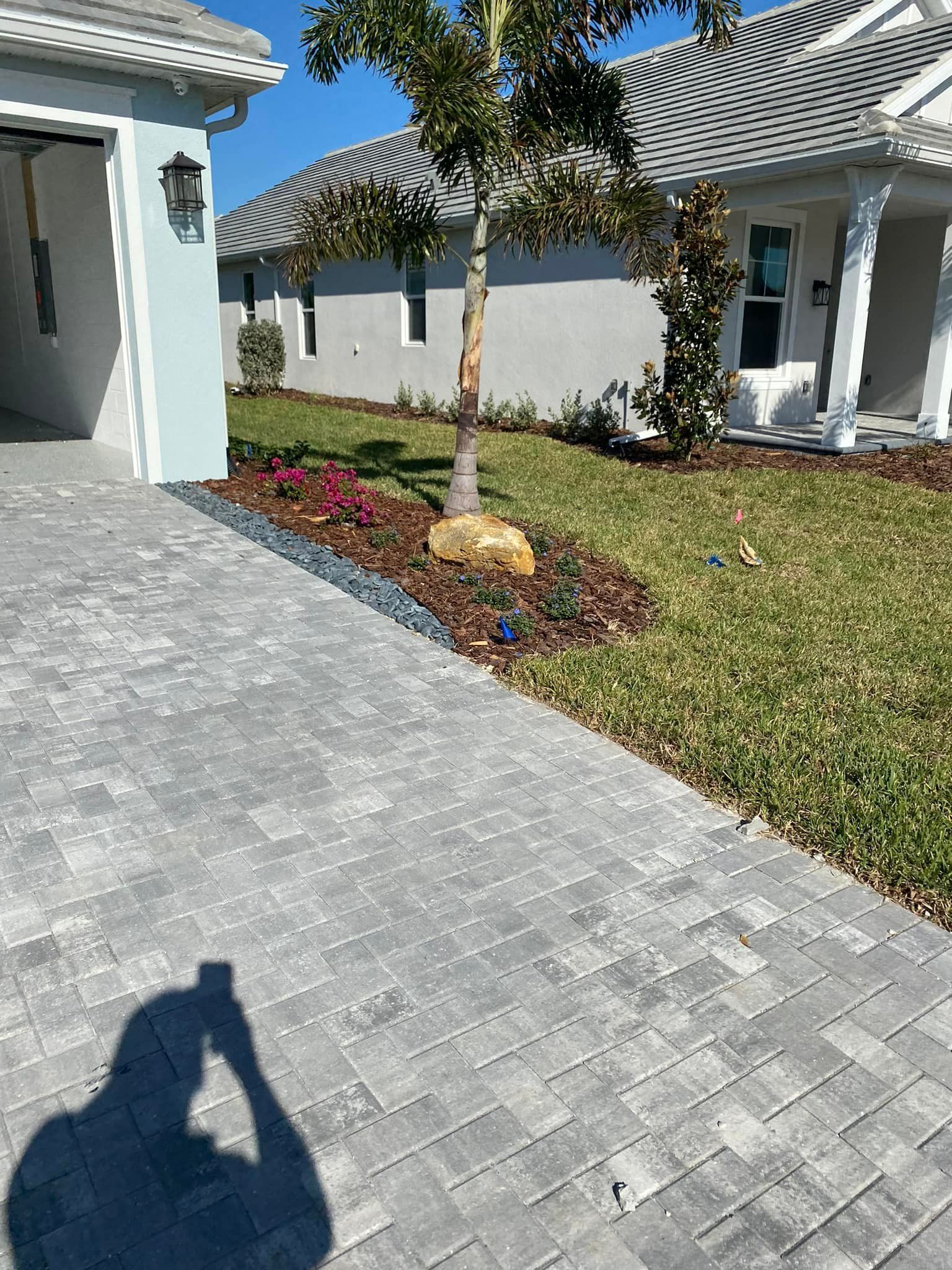 Gray brick driveway next to a grass lawn and flowerbed with a tree, houses in background.
