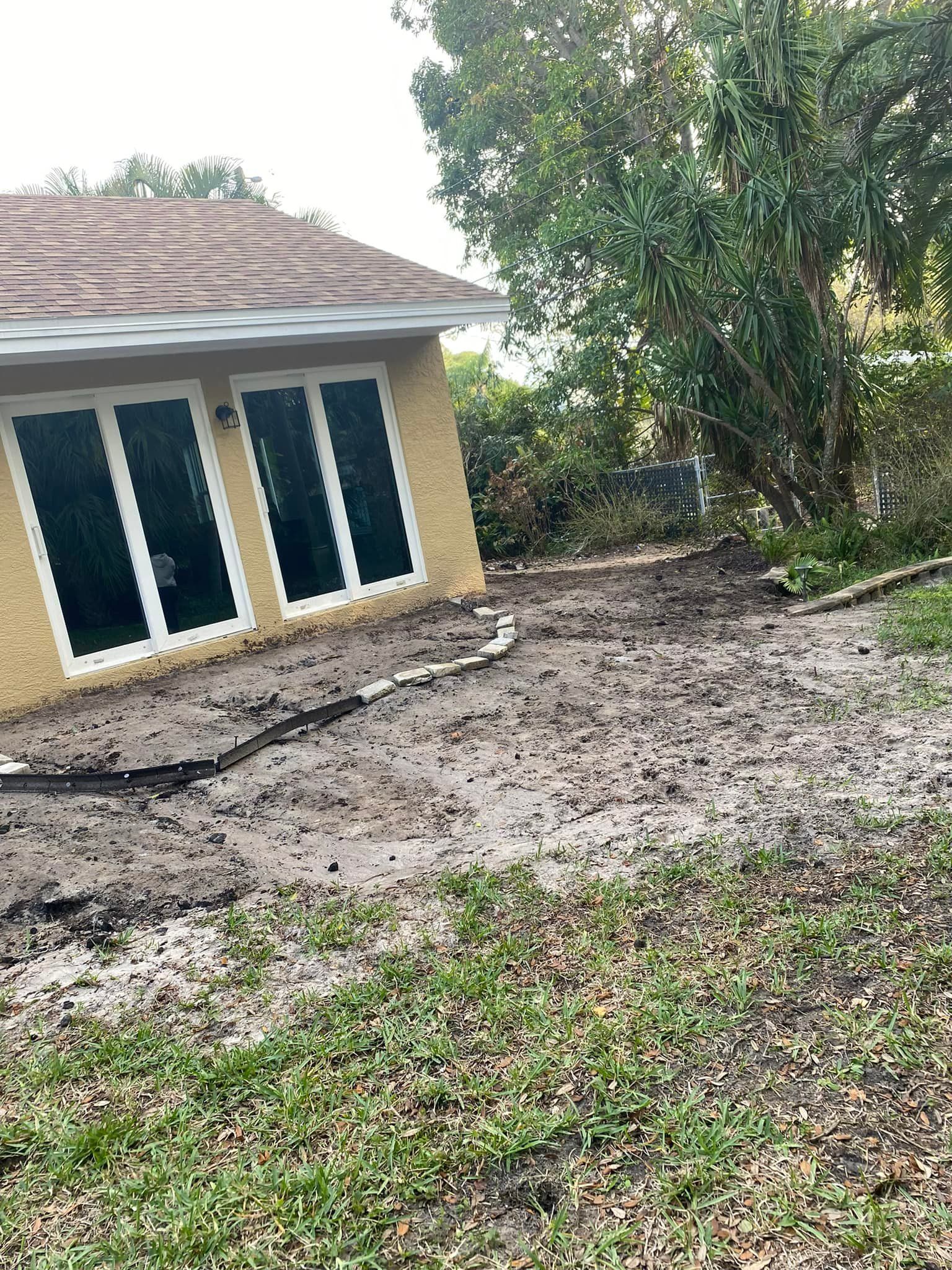 Backyard with muddy dirt area, possibly after landscaping. House with glass doors on the left, tree on the right.