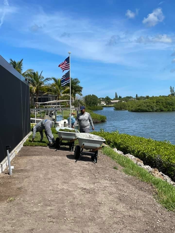 Workers laying sod along a waterfront path with an American flag on a sunny day.