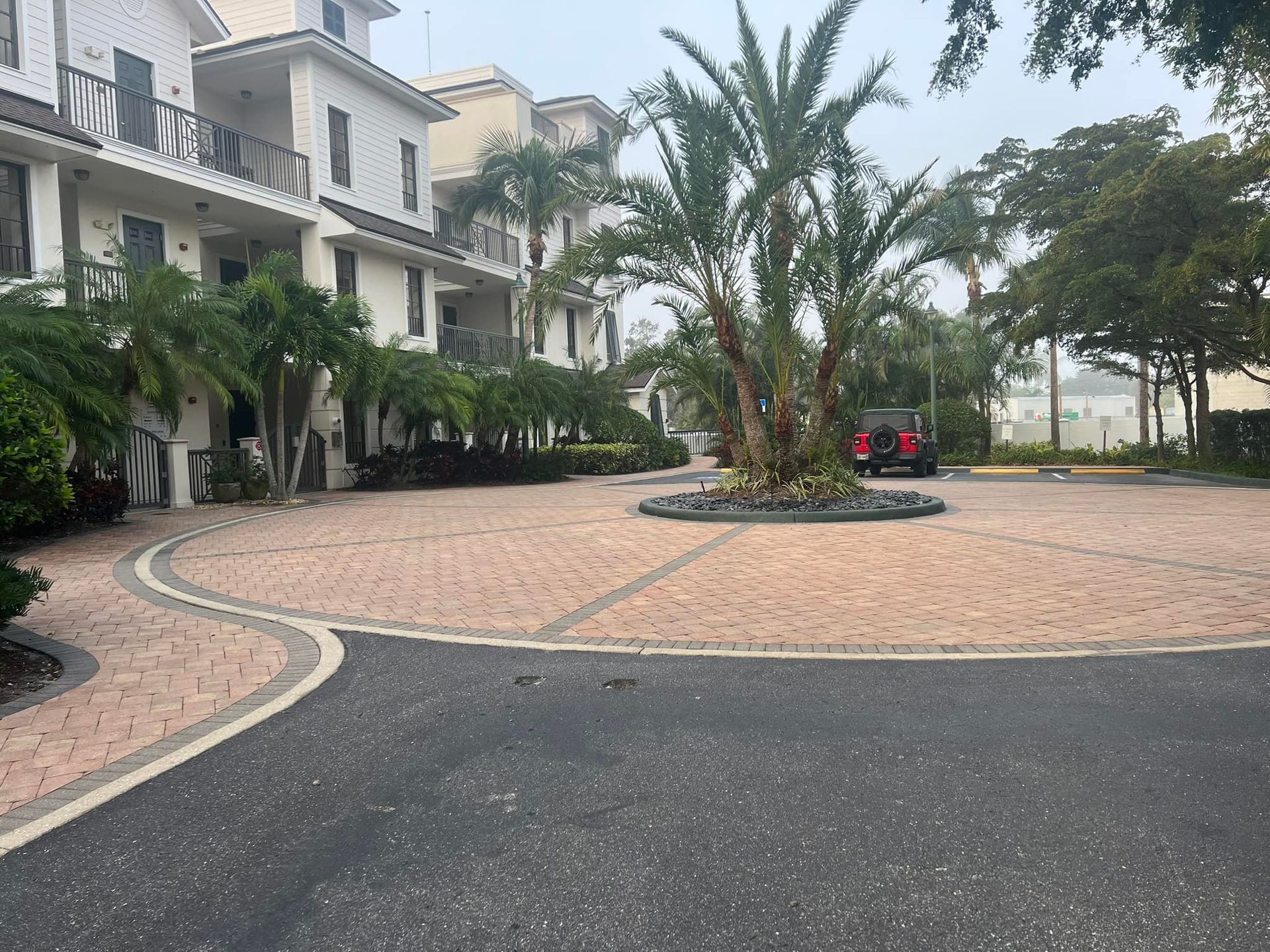 Brick paved driveway with palm tree island, in front of white multi-story buildings.