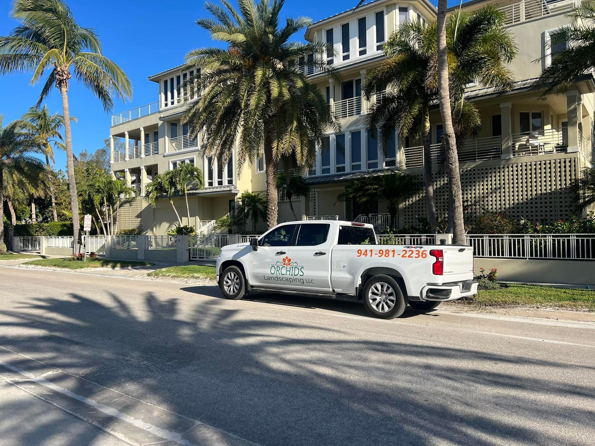 White pickup truck with company logo parked in front of a multi-story building and palm trees on a sunny day.