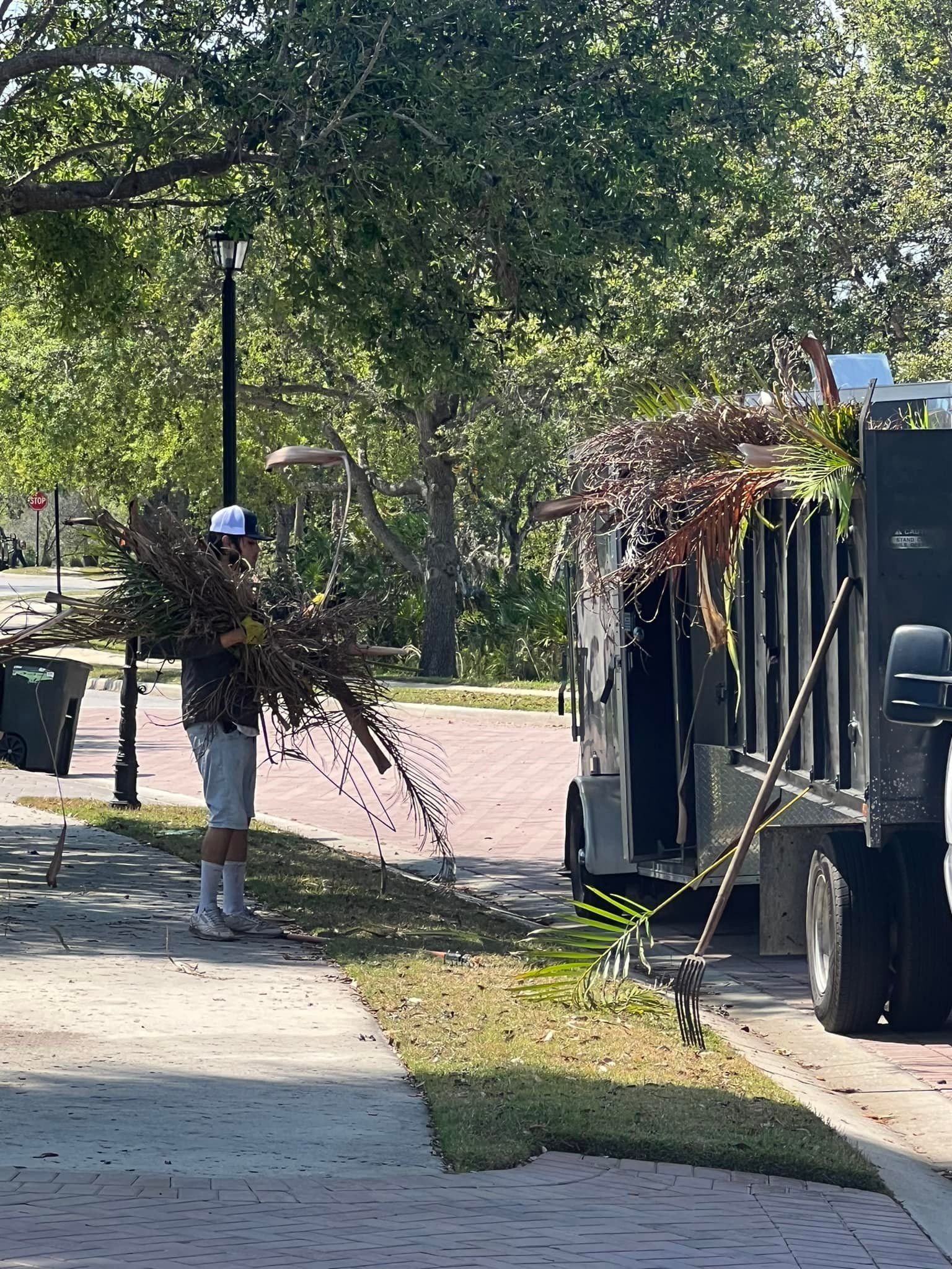 Man loads debris into a truck; he is wearing shorts and a hat, near a lawn.