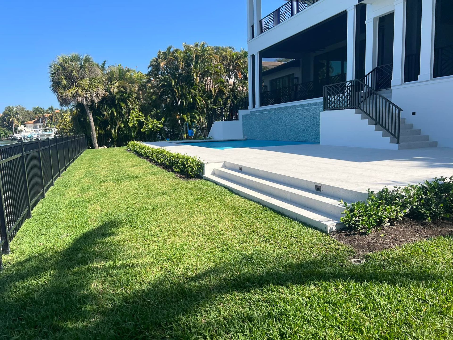 Lawn with a black fence, steps, a pool, and a white house with a balcony in a sunny outdoor setting.