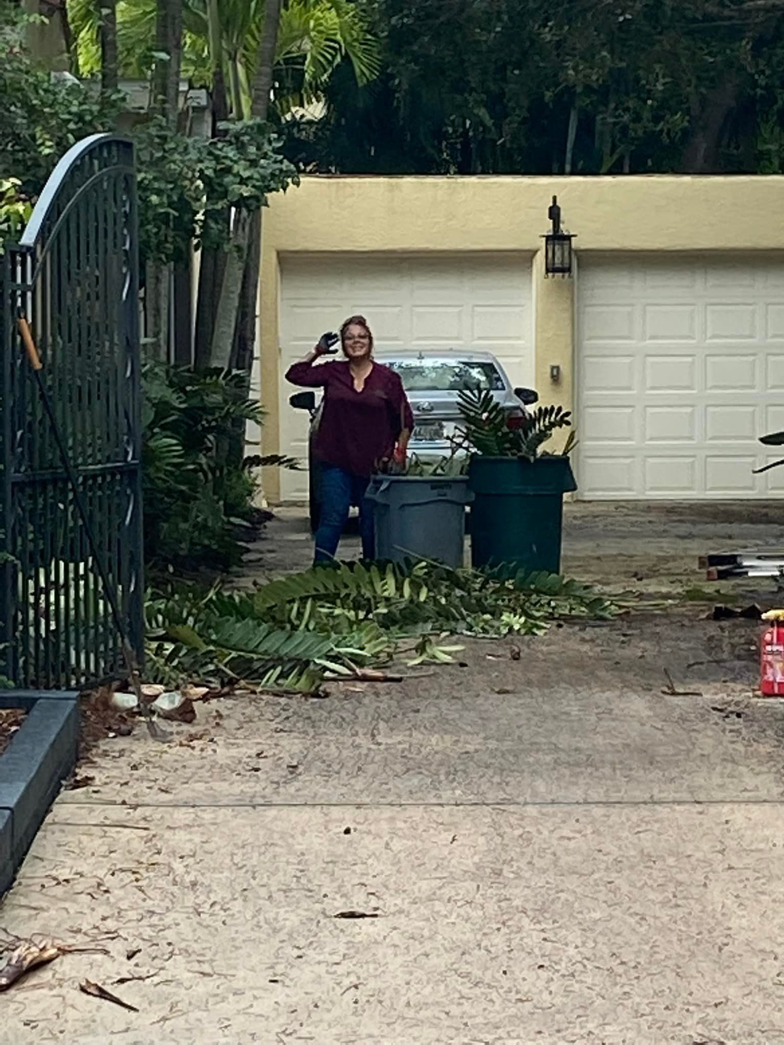 Woman standing in a driveway, gesturing, near trash cans and a car, with a garage in the background.