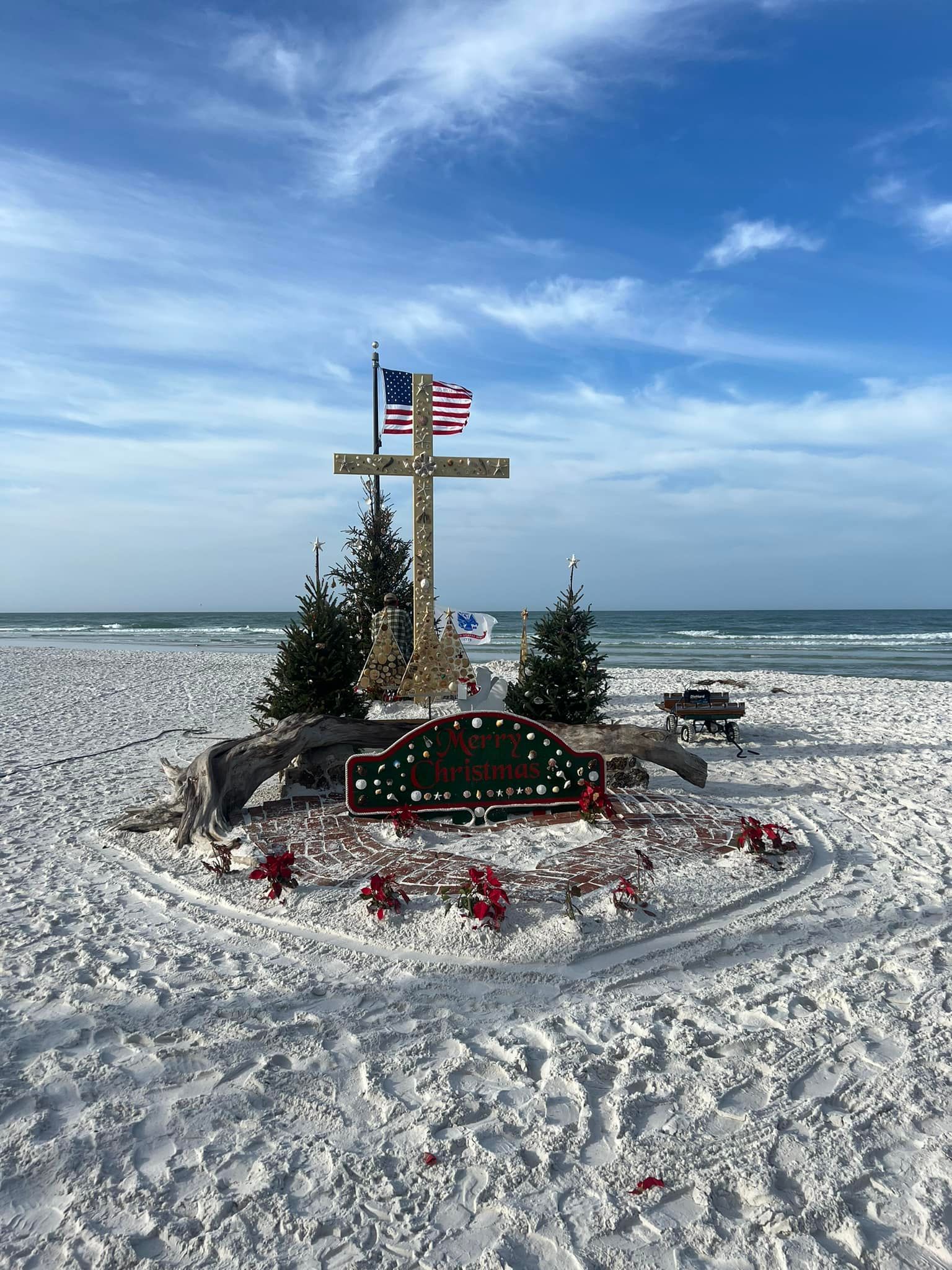 Beachfront memorial with cross, American flag, Christmas decorations, and two small trees.