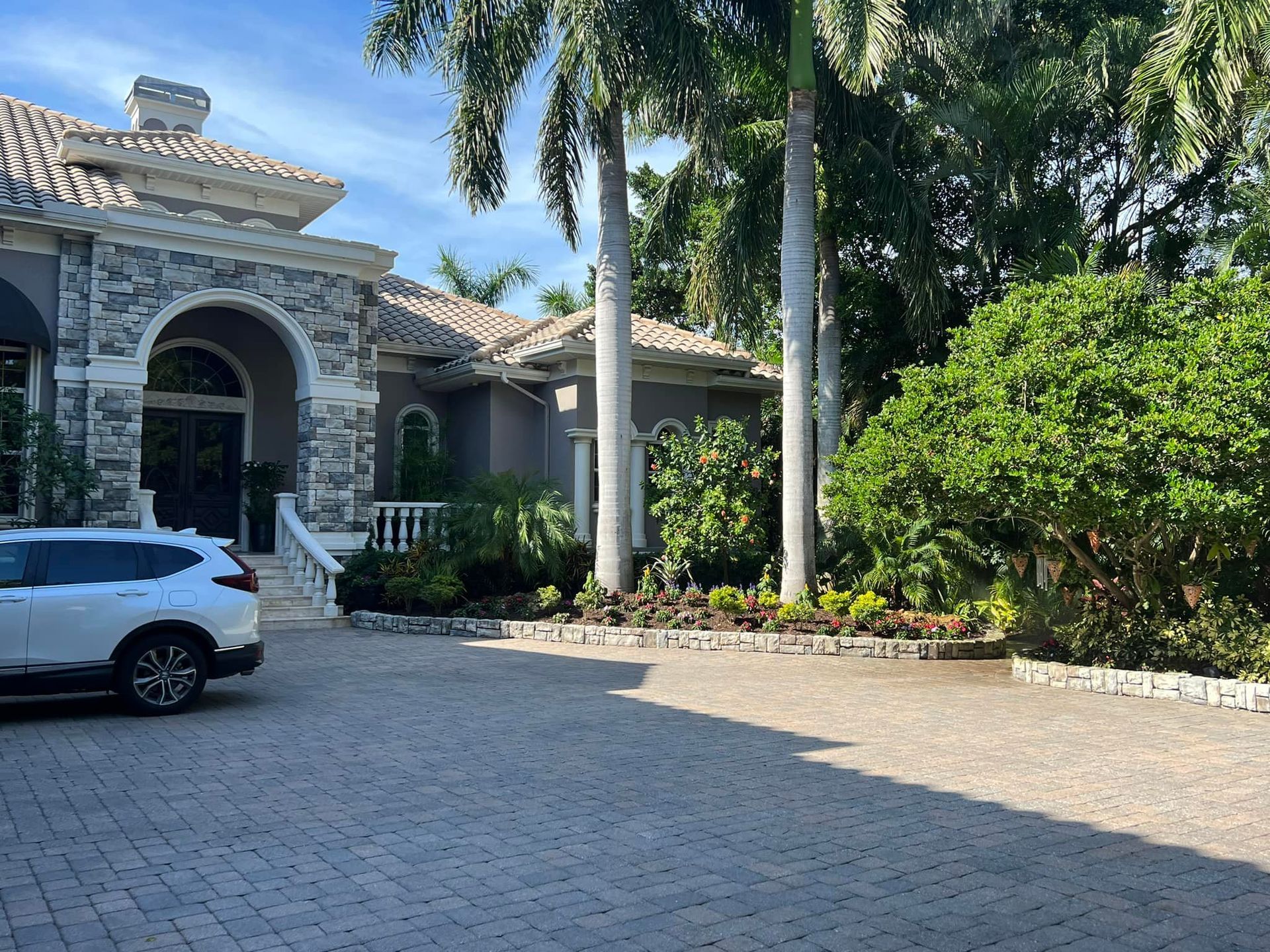 A large house with a stone facade, palm trees, and a brick driveway; a white SUV is parked.