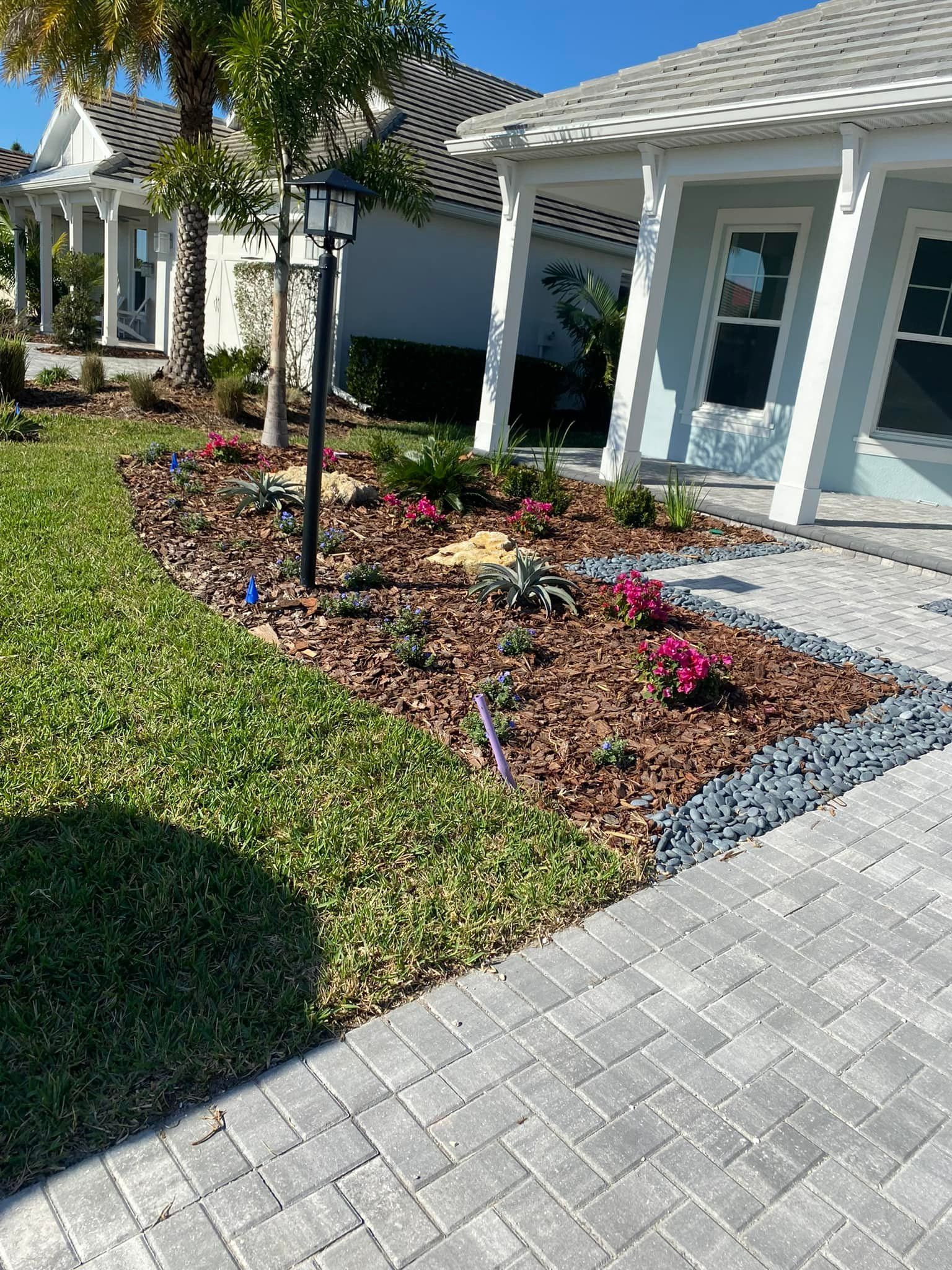 Flowerbed with pink blooms, mulch, dark stone border, next to a paved walkway and light blue house.