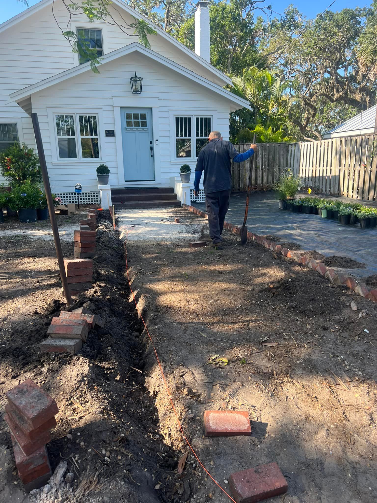 Person digging in a garden bed with red brick border in front of a white house with a blue door.