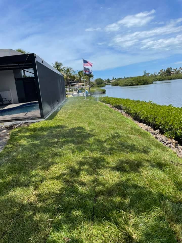 Lush green lawn being watered near a body of water, alongside a building and vegetation under a blue sky.