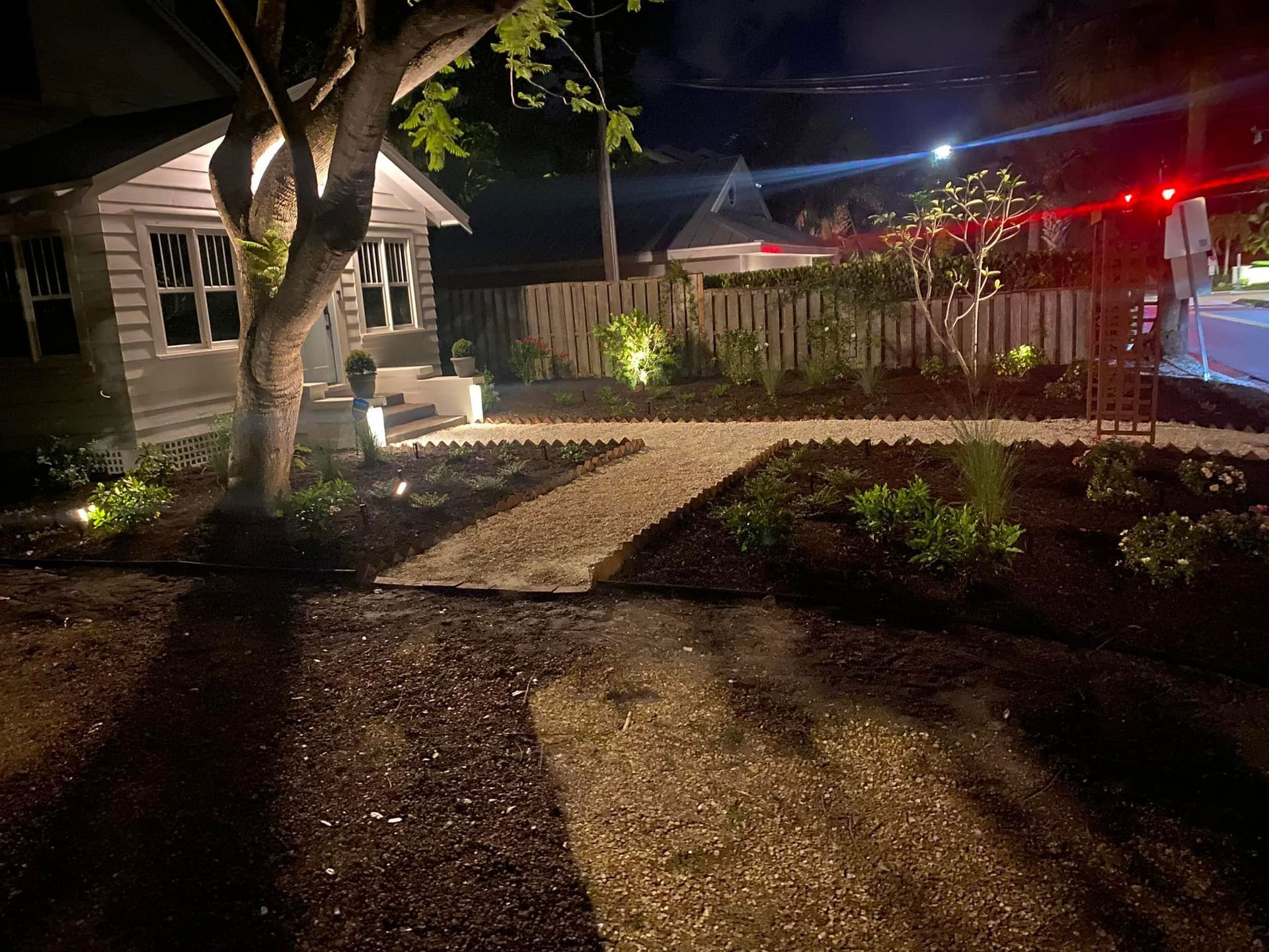 Night view of a yard with a lit pathway, a small house, and a garden. The path leads to the house, with lights.