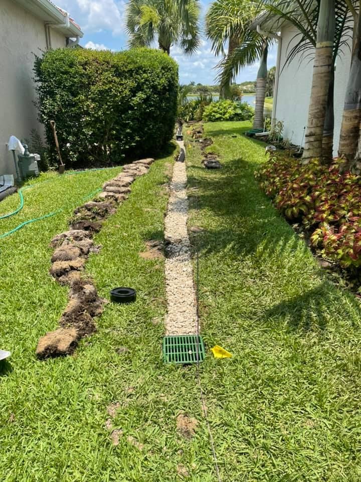 A trench with gravel and a drain in a grassy yard, next to a hedge and house.