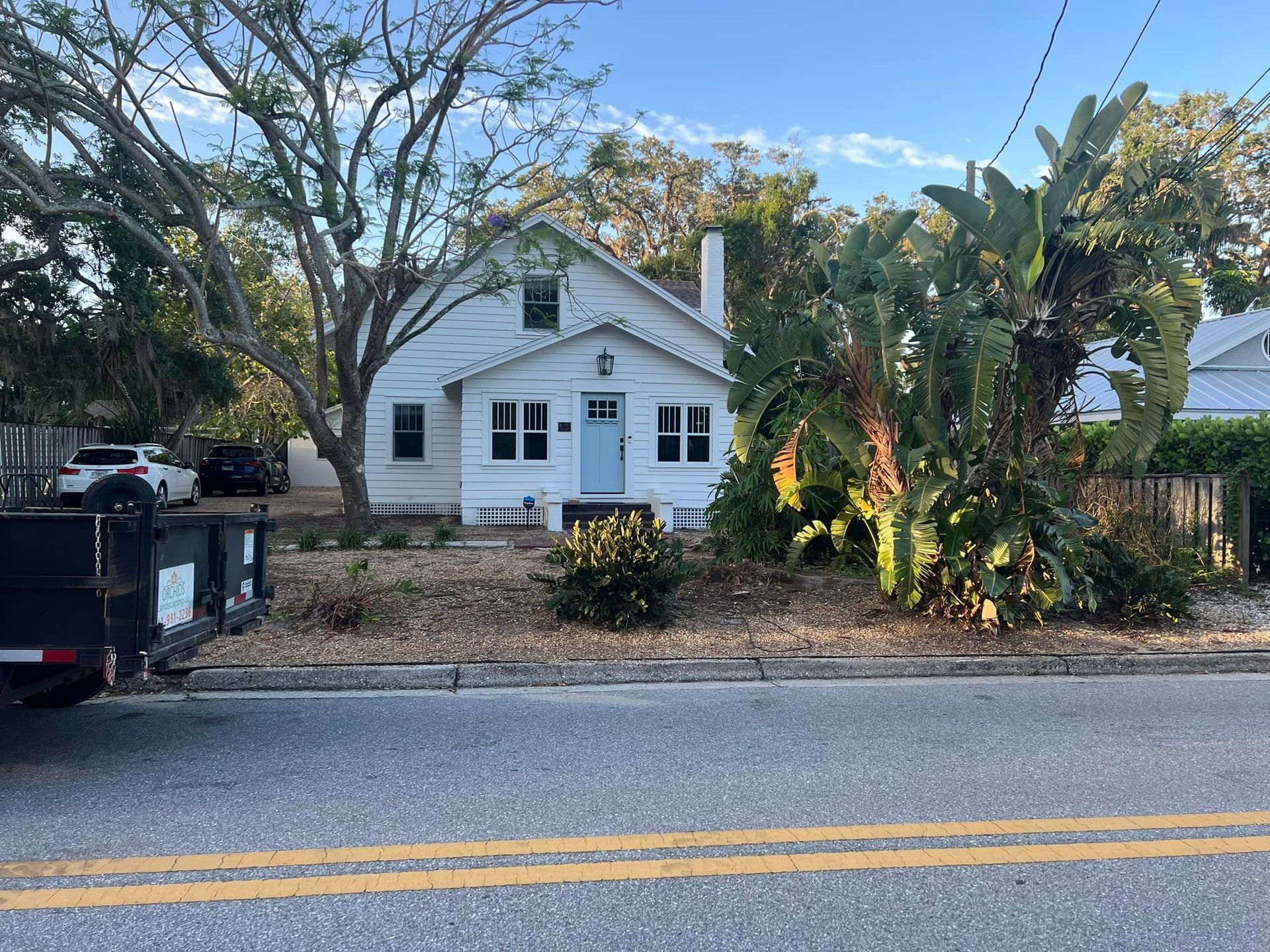 White house with blue door and windows, surrounded by trees and shrubs, on a roadside.